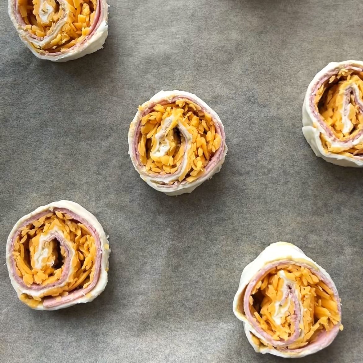 Five uncooked Ham and Cheese Pinwheels with visible cheese and ham layers are arranged on a baking tray lined with parchment paper.