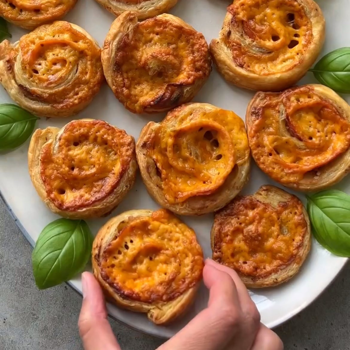 A hand places a Ham and Cheese Pinwheel onto a platter topped with more pinwheels and garnished with fresh basil.
