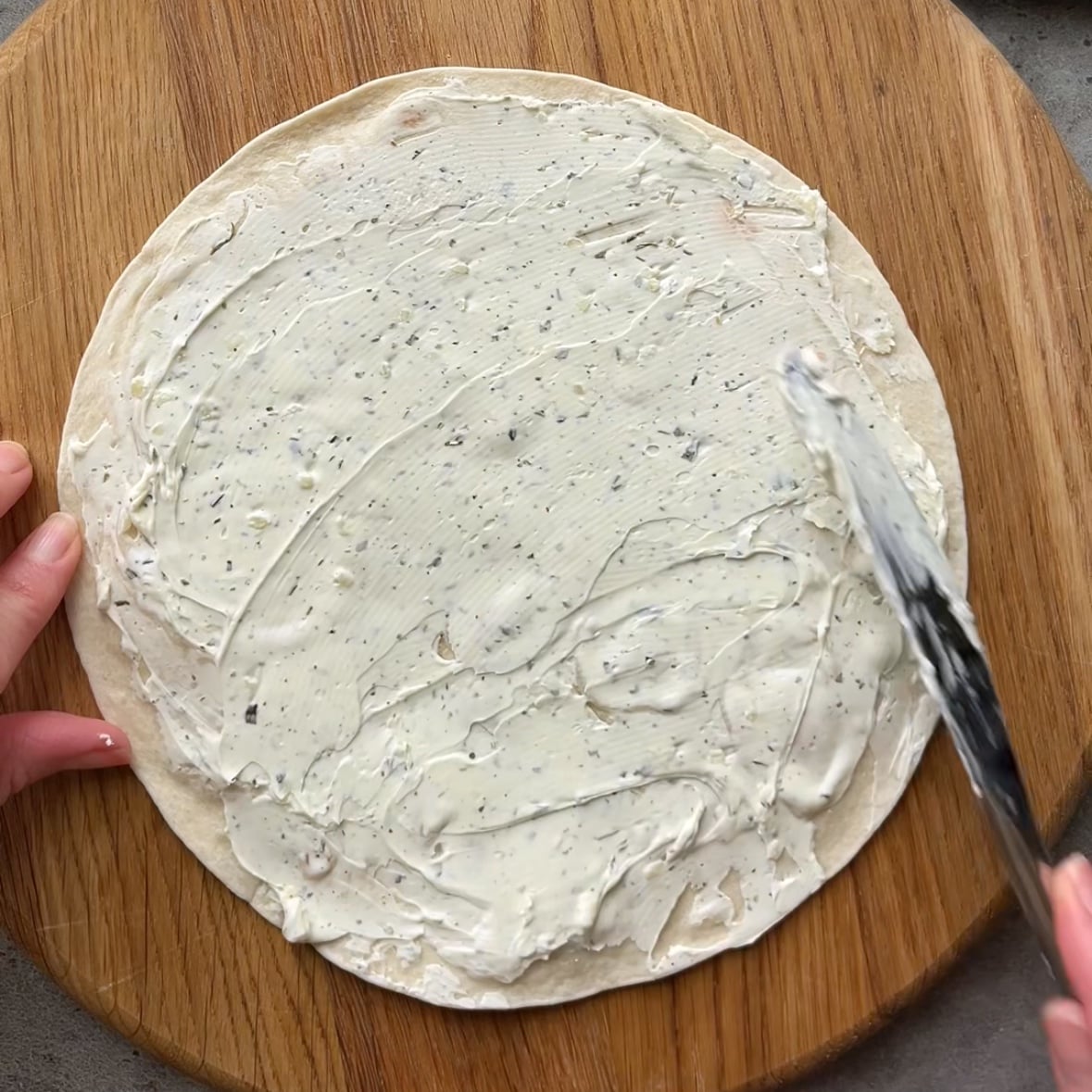 A plain white tortilla is being topped with garlic cream cheese.