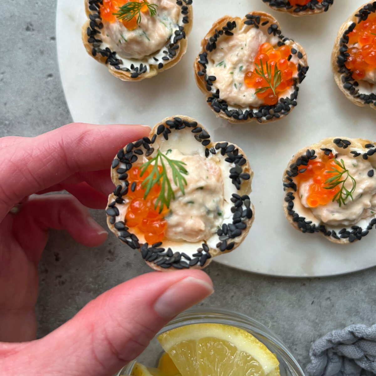 A hand holds a flower-shaped tortilla tart topped with cream cheese filling, orange fish roe, smoked salmon dip, black sesame seeds, and dill.