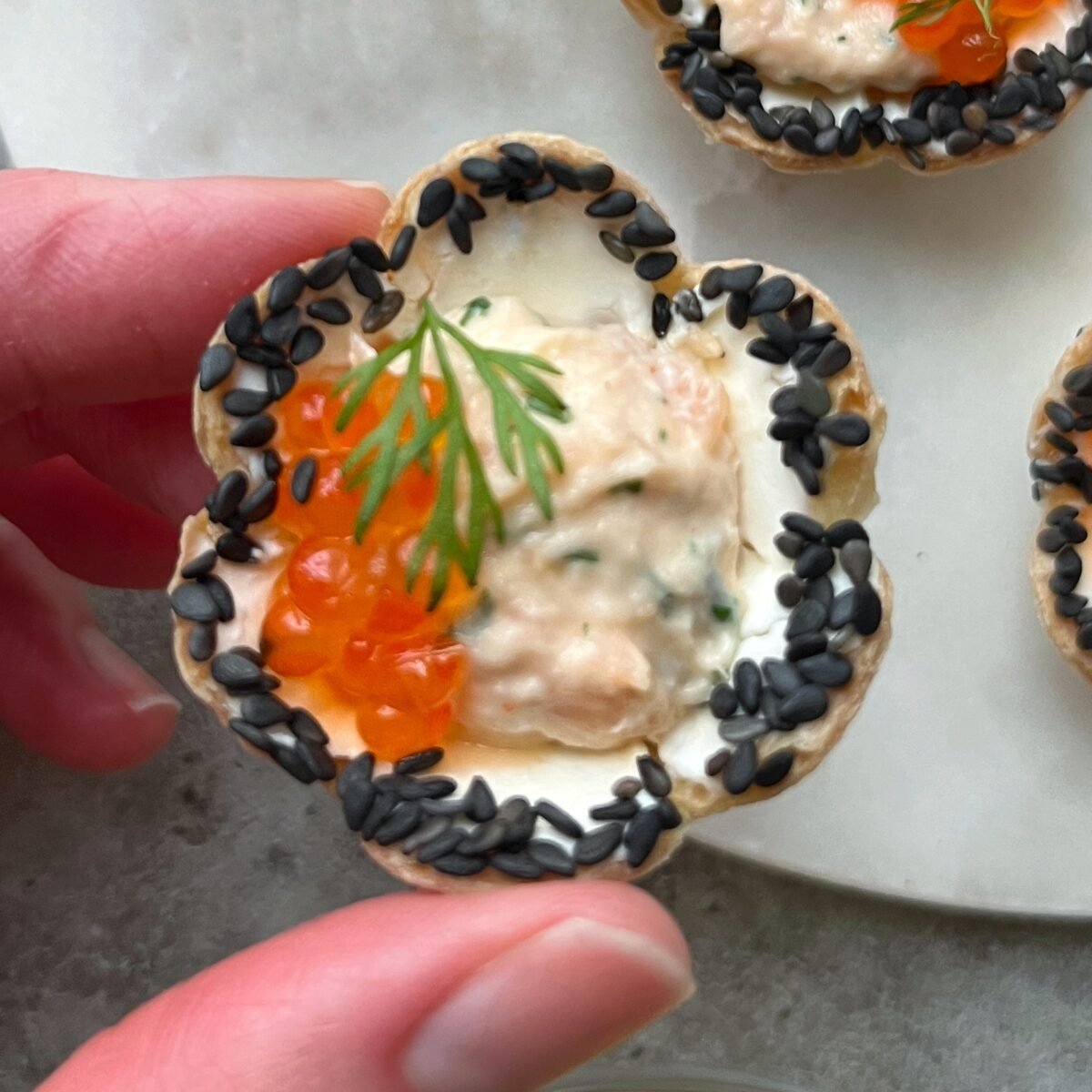 A hand holding a tortilla tart topped with a creamy seafood spread, orange fish roe, dill, and black sesame seeds along the edge.