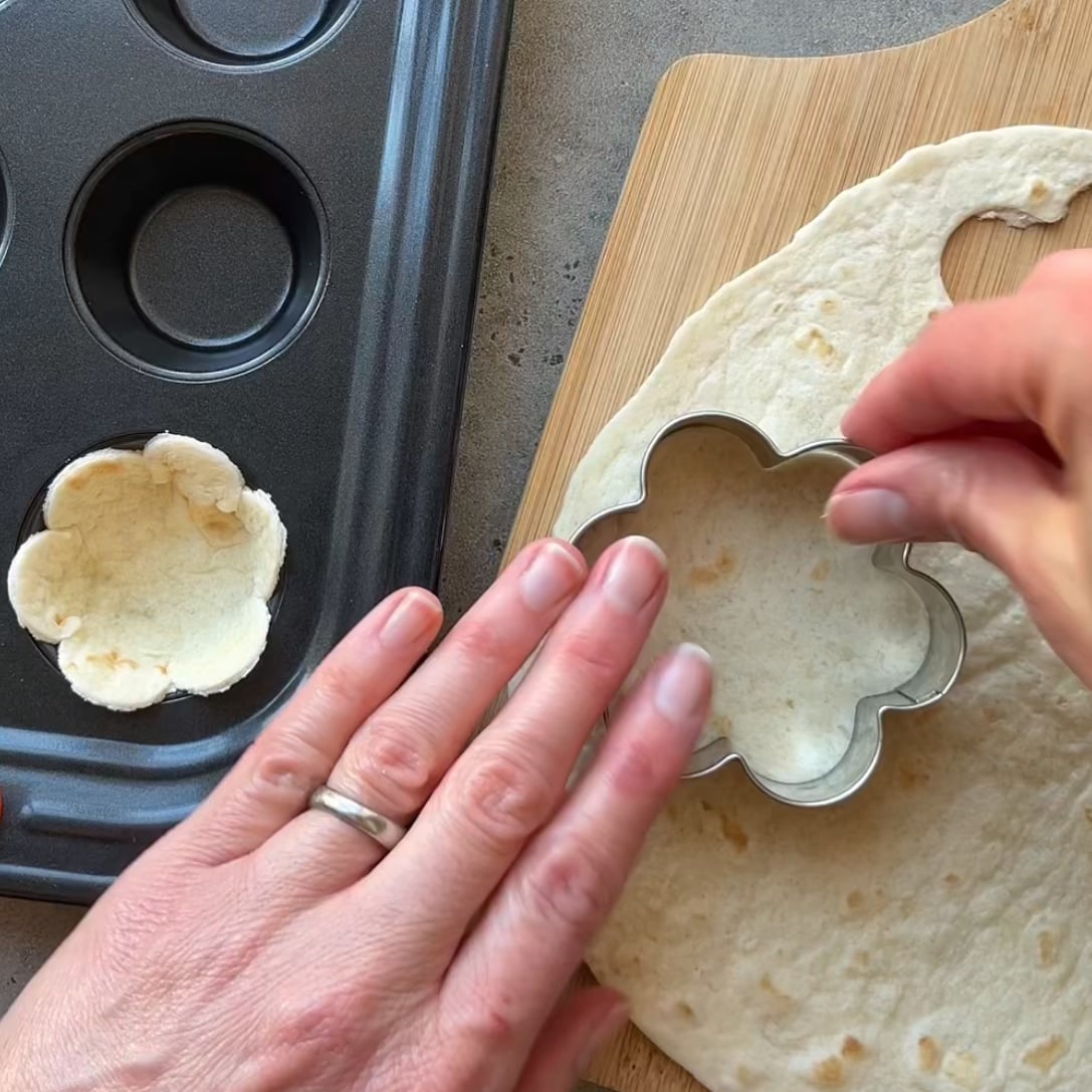 A person uses a flower-shaped cookie cutter on a tortilla next to a muffin tin and a wooden cutting board.