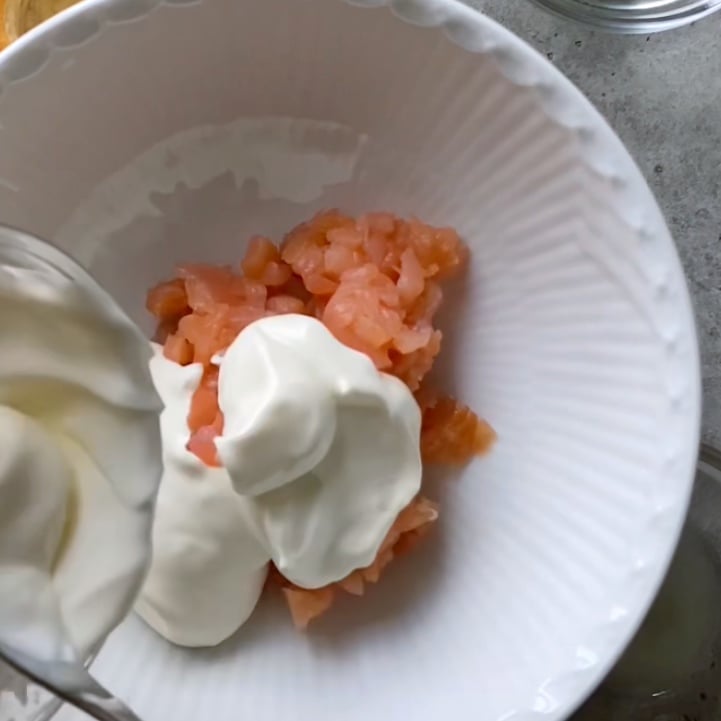 A white bowl contains raw diced smoked salmon and a dollop of sour cream, being added with a spoon.