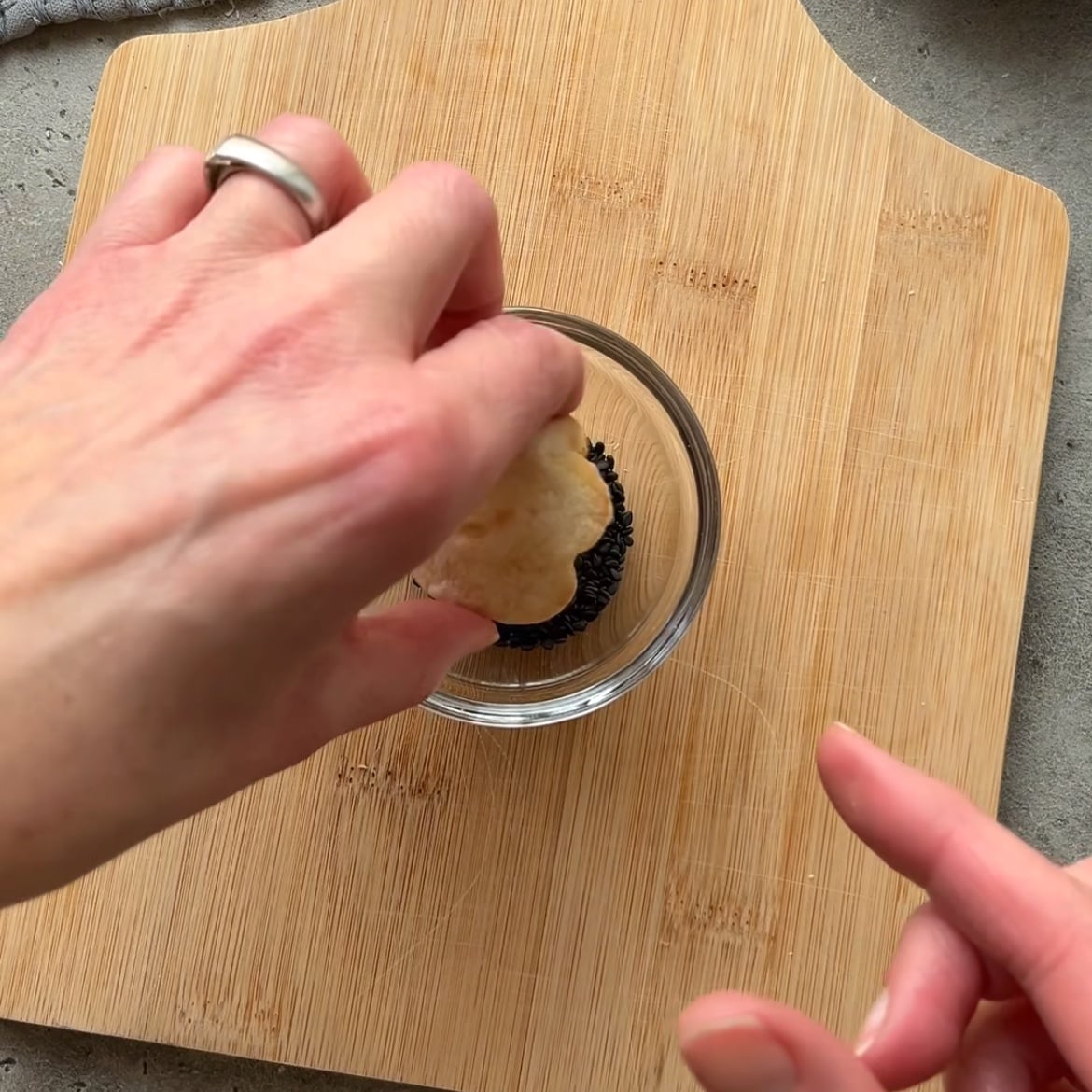 A hand dips a round cracker into a small glass bowl filled with a dark spread, placed on a wooden cutting board.