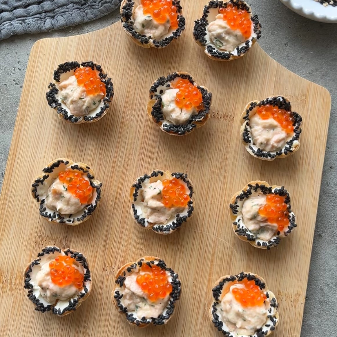 Twelve bite-sized Smoked Salmon Dip Bites with cream filling, black sesame seeds on the edges, and orange fish roe topping, arranged on a wooden board.