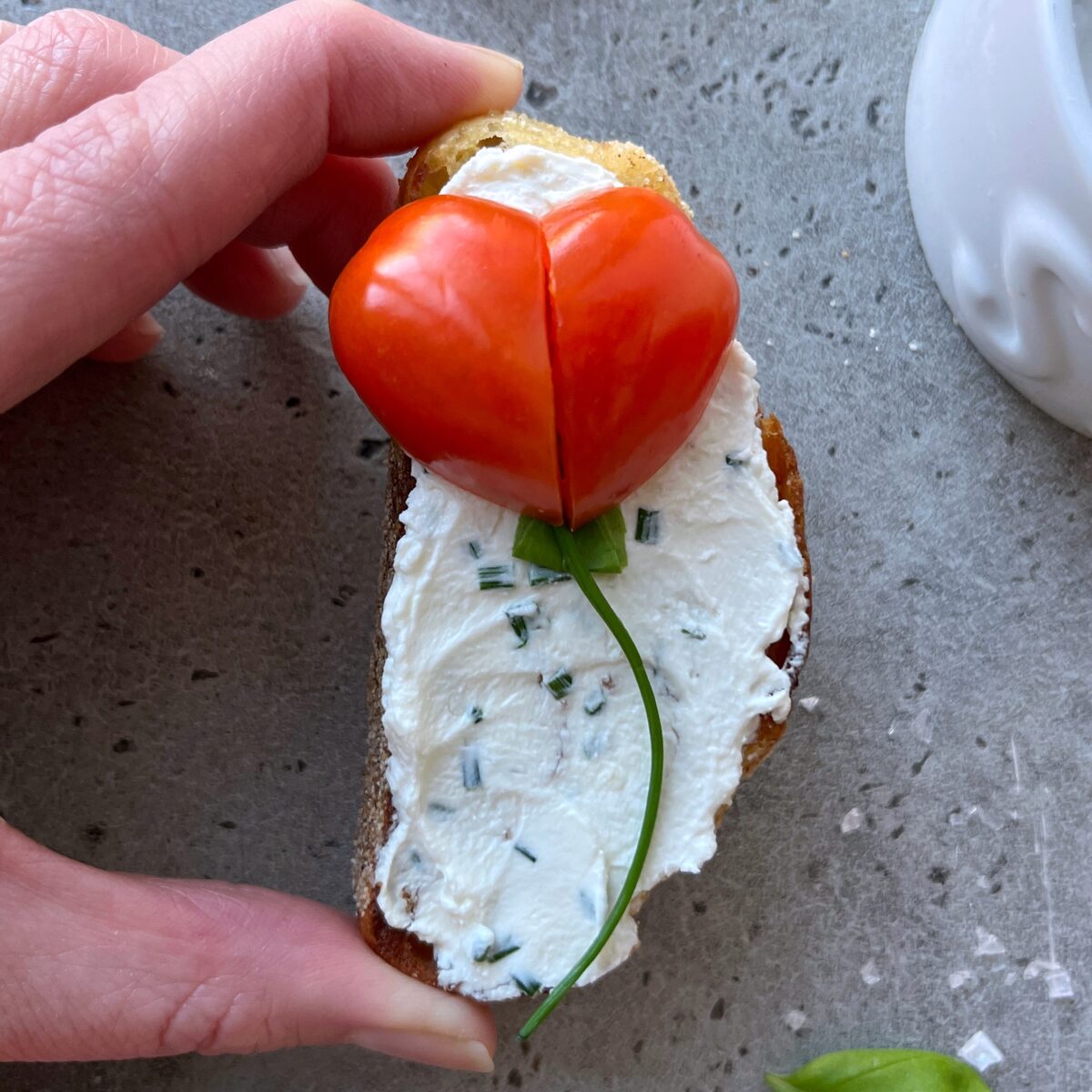 A hand holds a slice of bread topped with cream cheese, chopped chives, and a heart-shaped tomato with a green stem.