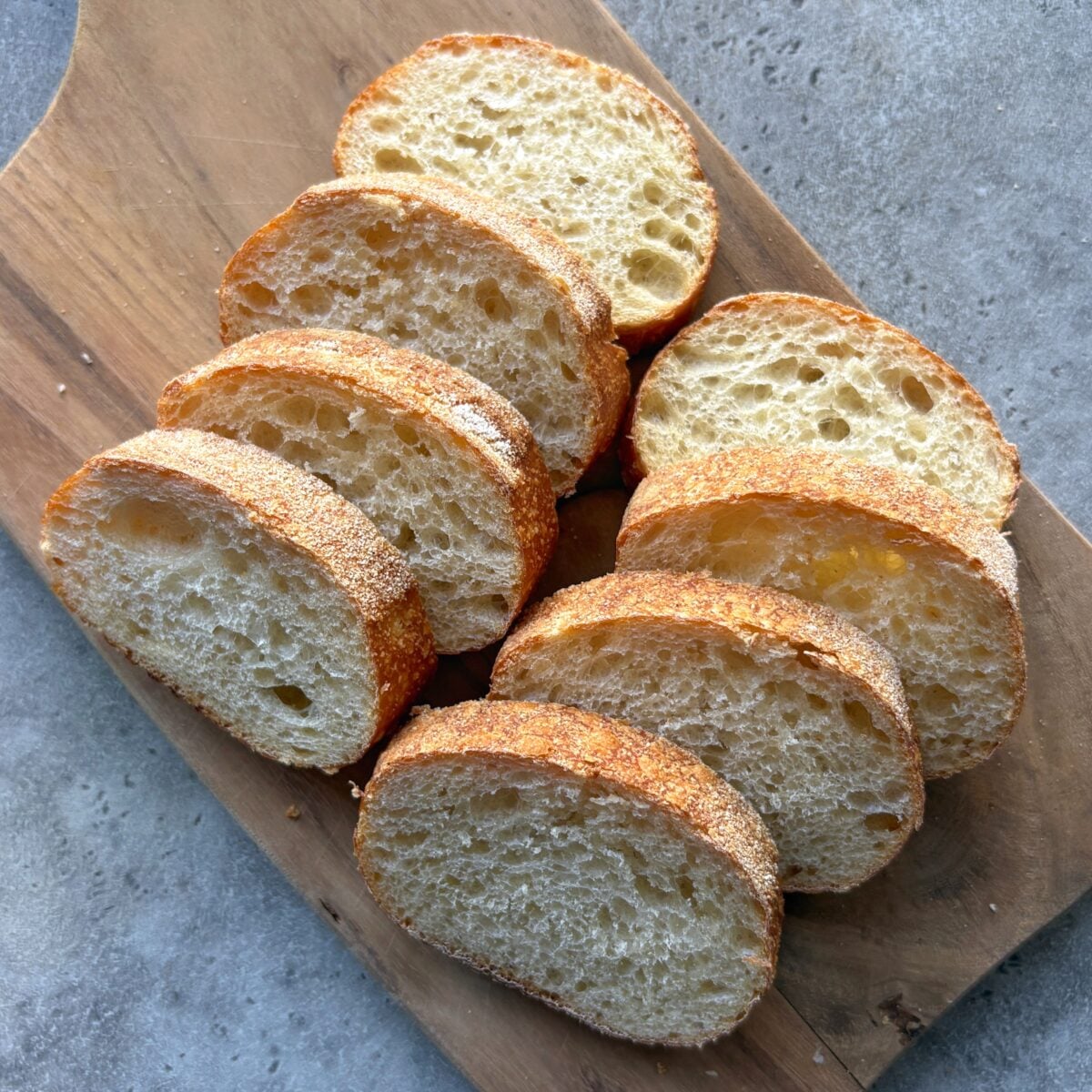 Eight slices of rustic bread are arranged on a wooden cutting board, ready to be transformed into delicious Tomato Crostini, and placed on a gray countertop.
