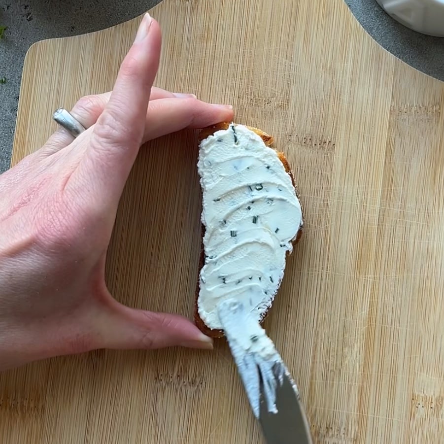 A hand spreads herb cream cheese on a slice of bread using a knife.