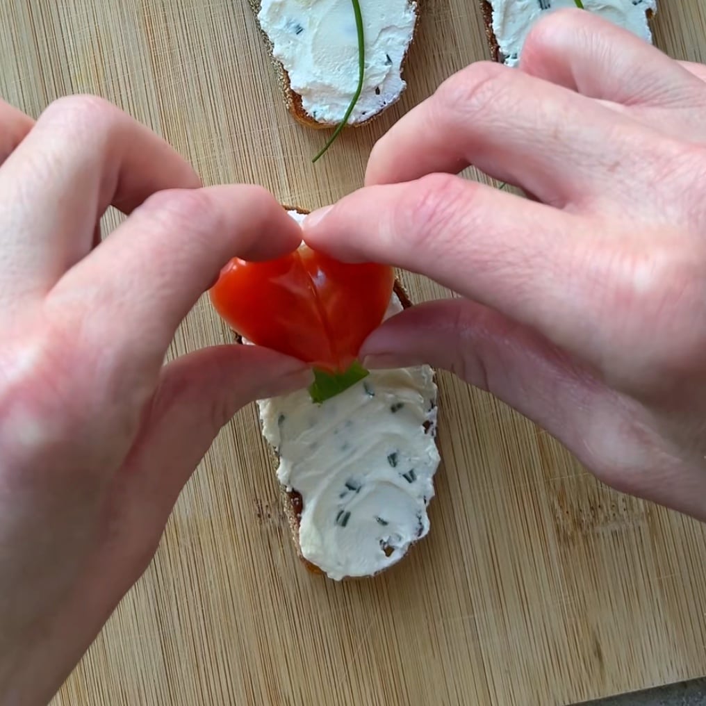 Two hands arrange a heart shape made with  sliced tomato on bread topped with cream cheese and herbs on a wooden cutting board.