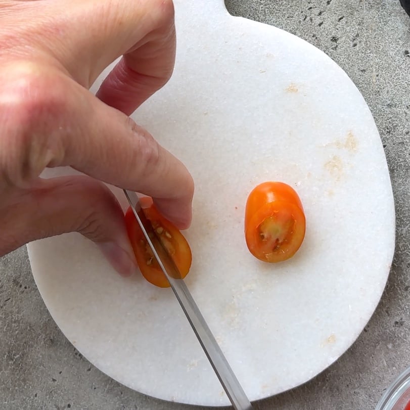 A hand is slicing a small grape tomato in half on a white cutting board with a knife.