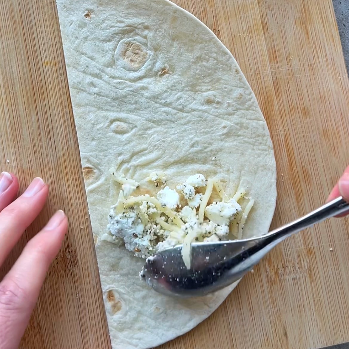 A person spreads shredded cheese onto half of a flour tortilla with a spoon on a wooden cutting board.