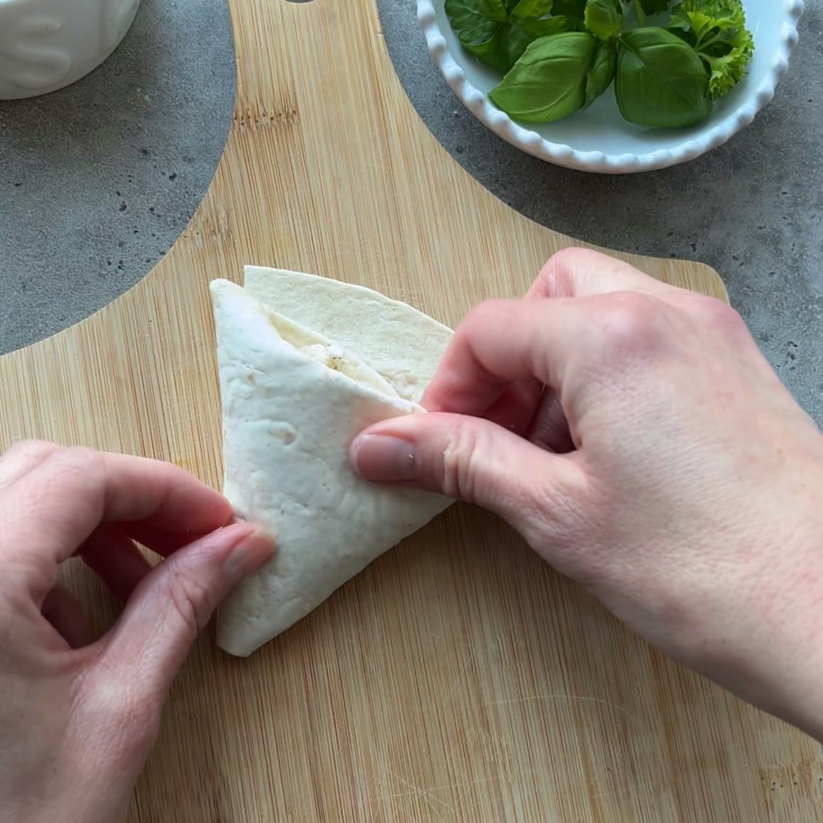 Two hands folding Tortilla Pockets with Feta on a wooden cutting board, with a small bowl of fresh basil leaves nearby.