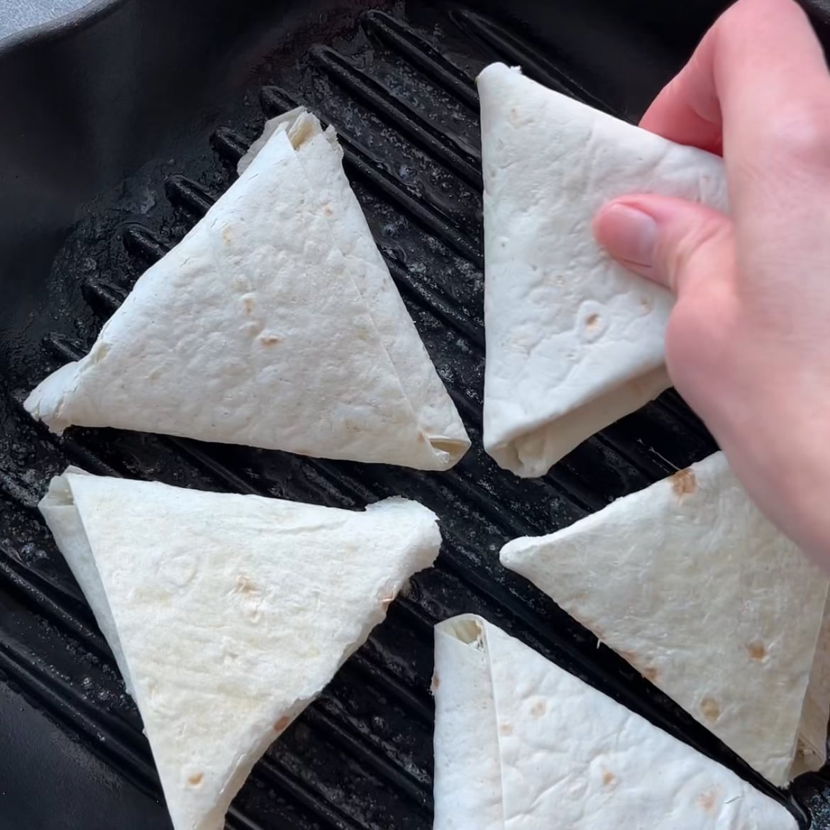 A hand places a folded tortilla pockets onto a grill pan alongside four other similar tortilla triangles.