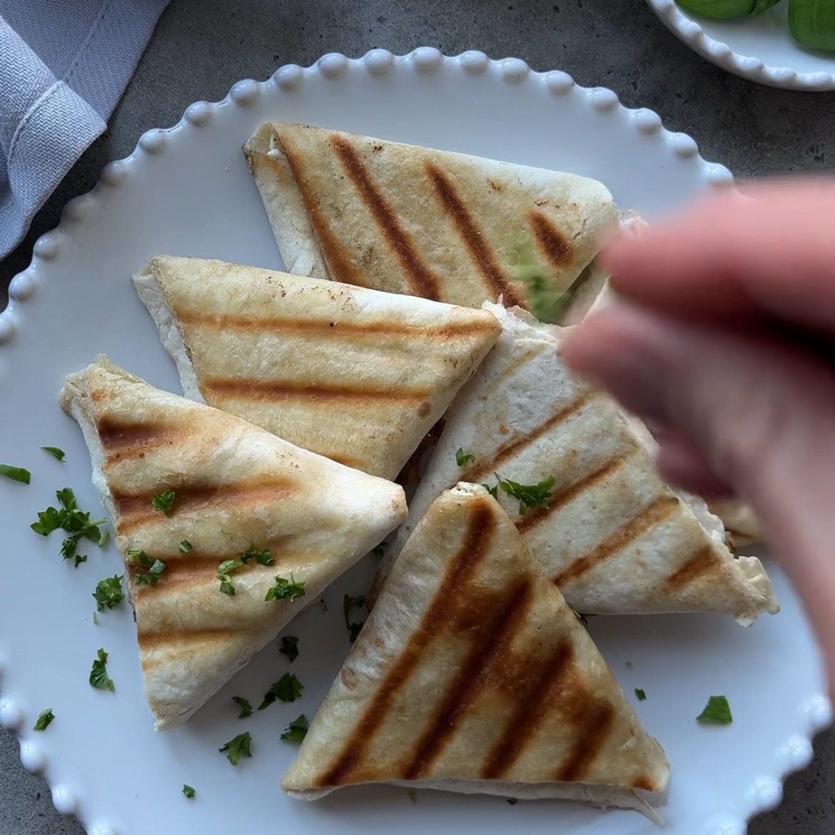 Six grilled Tortilla Pockets with Feta are arranged in triangles on a white plate, while a hand sprinkles chopped parsley on top.
