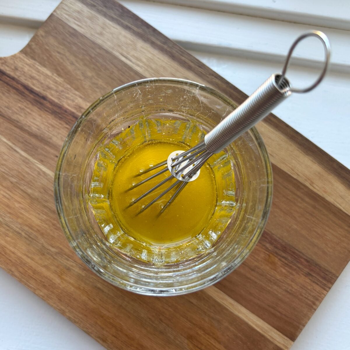 A small glass bowl with yellow dressing and a metal whisk sits on a wooden cutting board, ready to be used as the dressing for a fresh Tuna Cottage Cheese Salad.