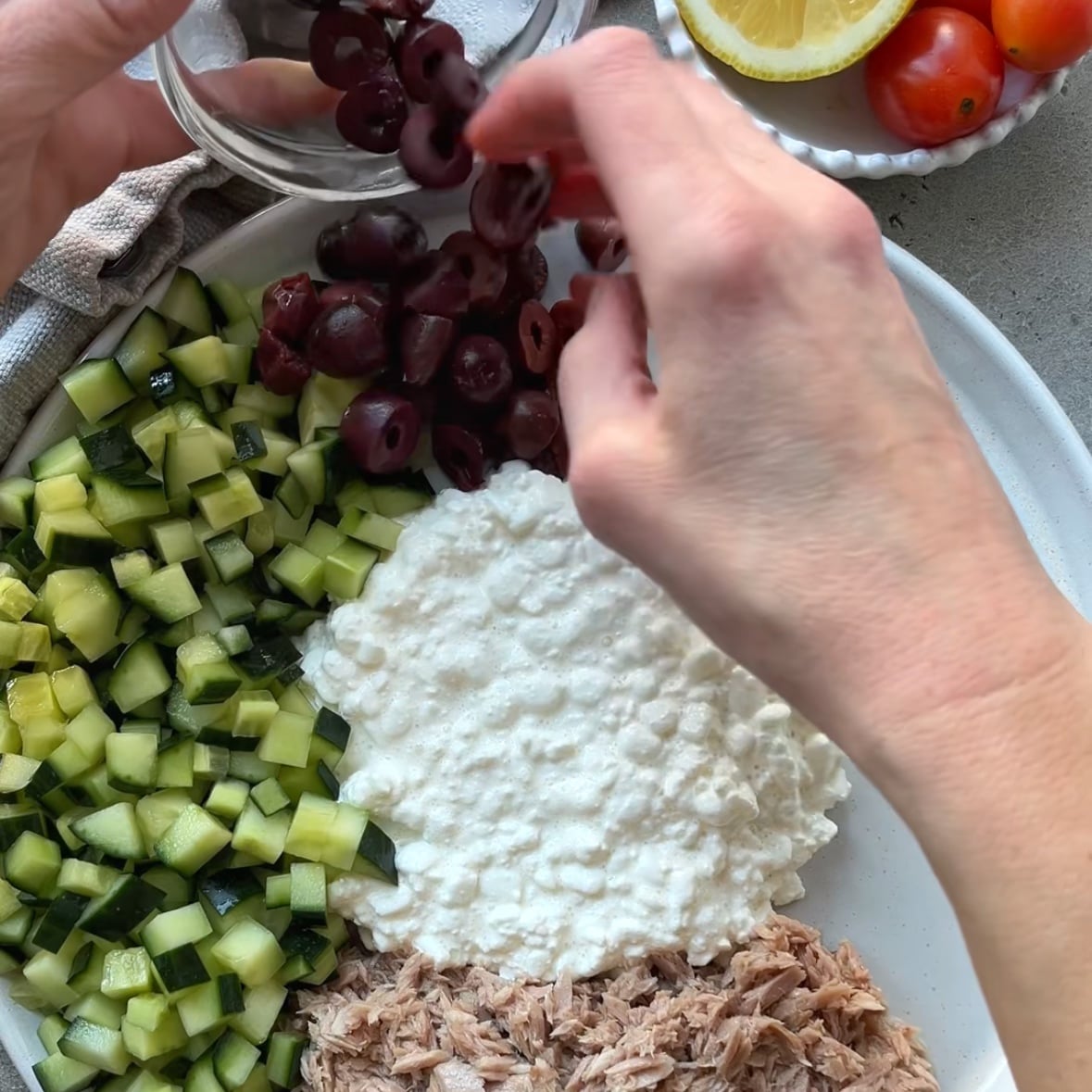 A person prepares a Tuna Cottage Cheese Salad by adding sliced kalamata olives to a plate with diced cucumber, cottage cheese, and tuna. A bowl of cherry tomatoes and a lemon wedge is nearby.