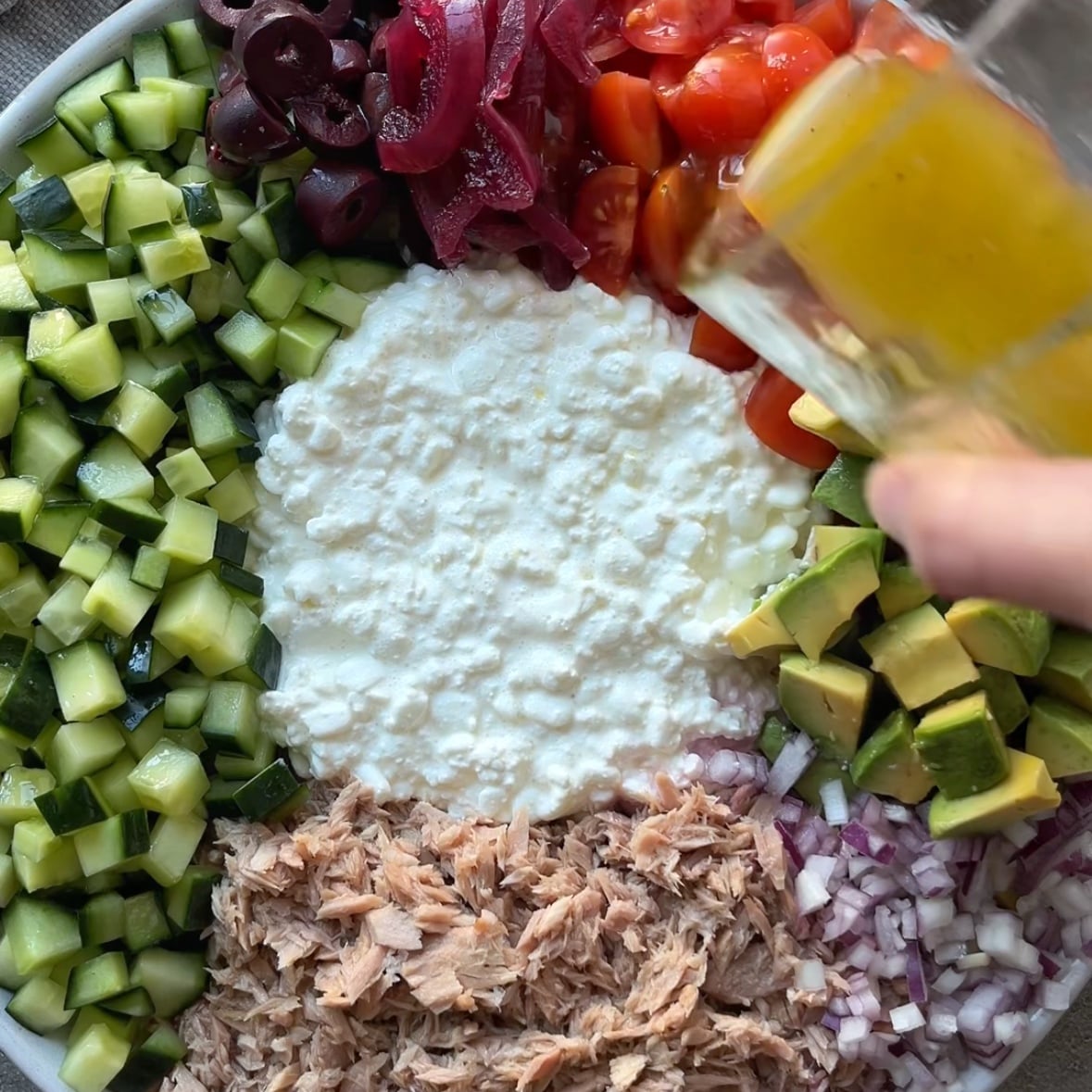 A bowl features cottage cheese, diced cucumber, cherry tomatoes, avocado, onions, olives, pickled onions, and chunks of tuna. A hand pours a dressing on top.