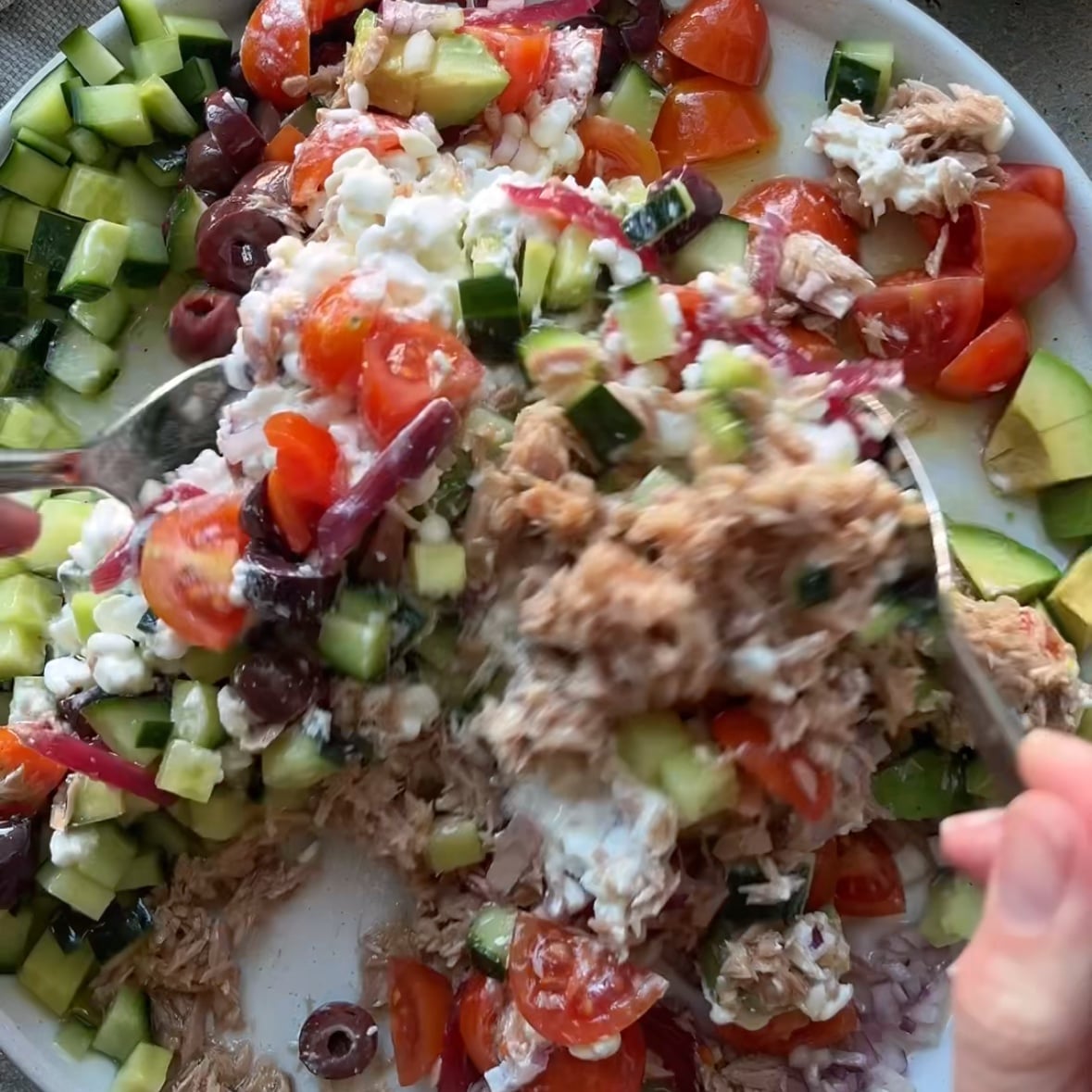 A person mixes a Tuna Cottage Cheese Salad with cherry tomatoes, cucumbers, olives, and red onions on a white plate using a fork and spoon.