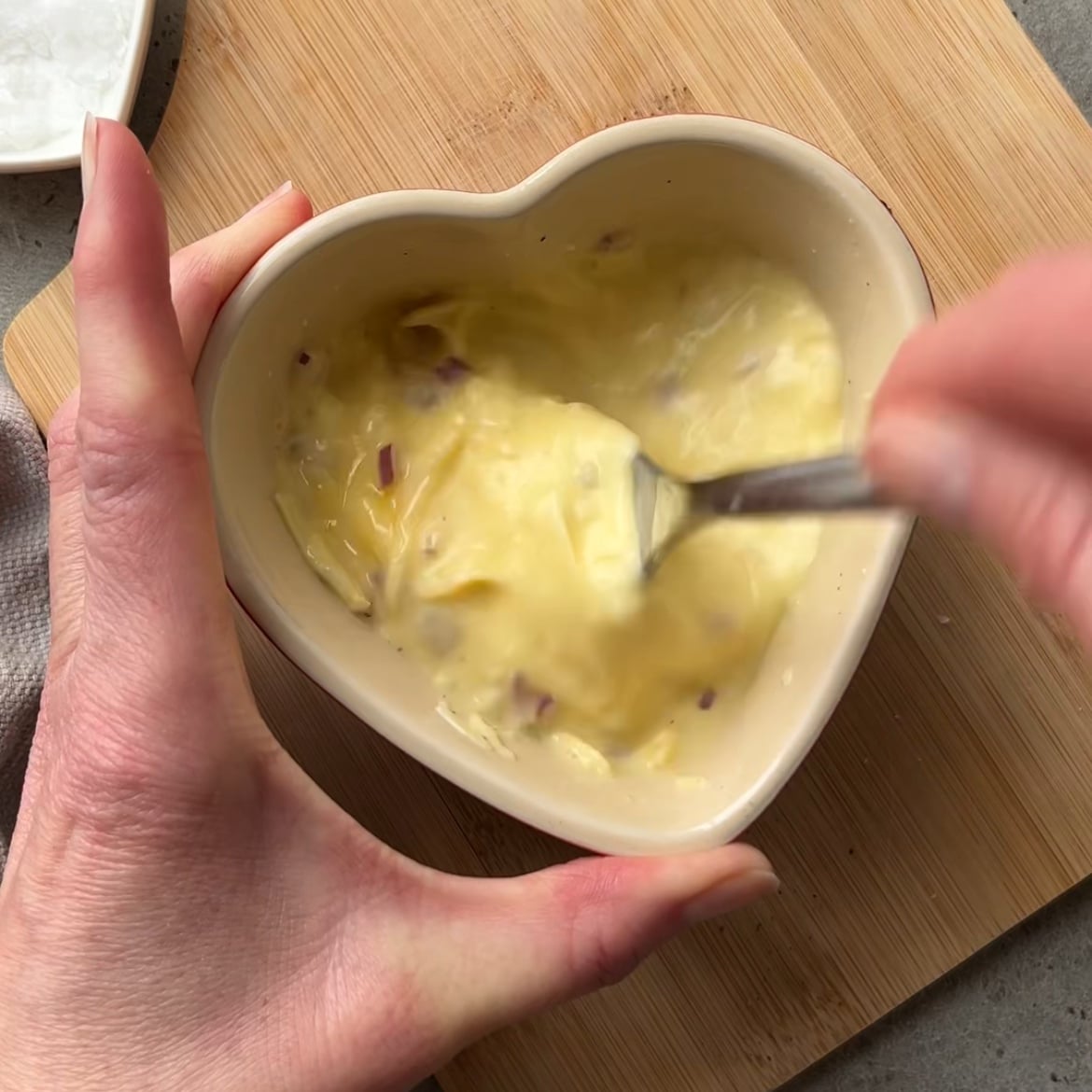 A person stirs a creamy mixture with small chunks for Baked Egg Tortilla Bites in a heart-shaped bowl using a fork, placed on a wooden board.