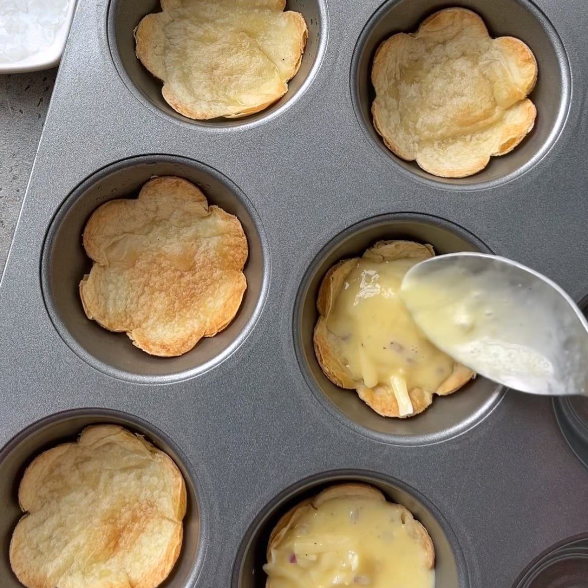 A muffin tin holds Baked Egg Tortilla Bites; a spoon is filling one shell with a creamy yellow sauce.