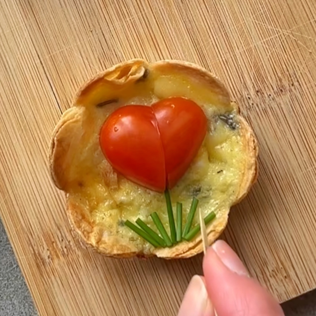 A small Baked Egg Tortilla Bite with egg and melted cheese is topped with a heart-shaped cherry tomato and chives.