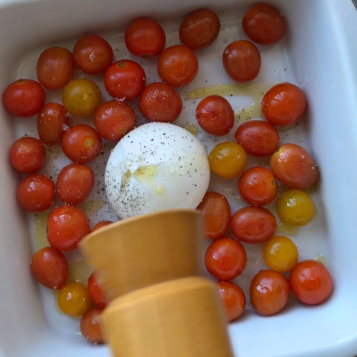 A white baking dish with cherry tomatoes and a ball of burrata cheese is being seasoned with freshly ground pepper.