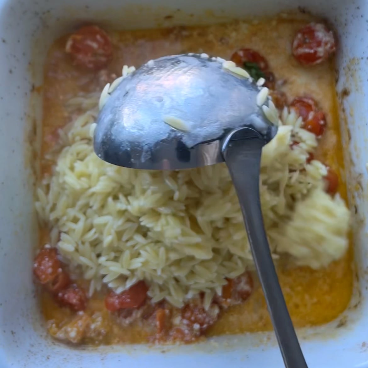 A metal ladle holds cooked orzo over a baking dish containing orzo, cherry tomatoes, and a creamy orange sauce.