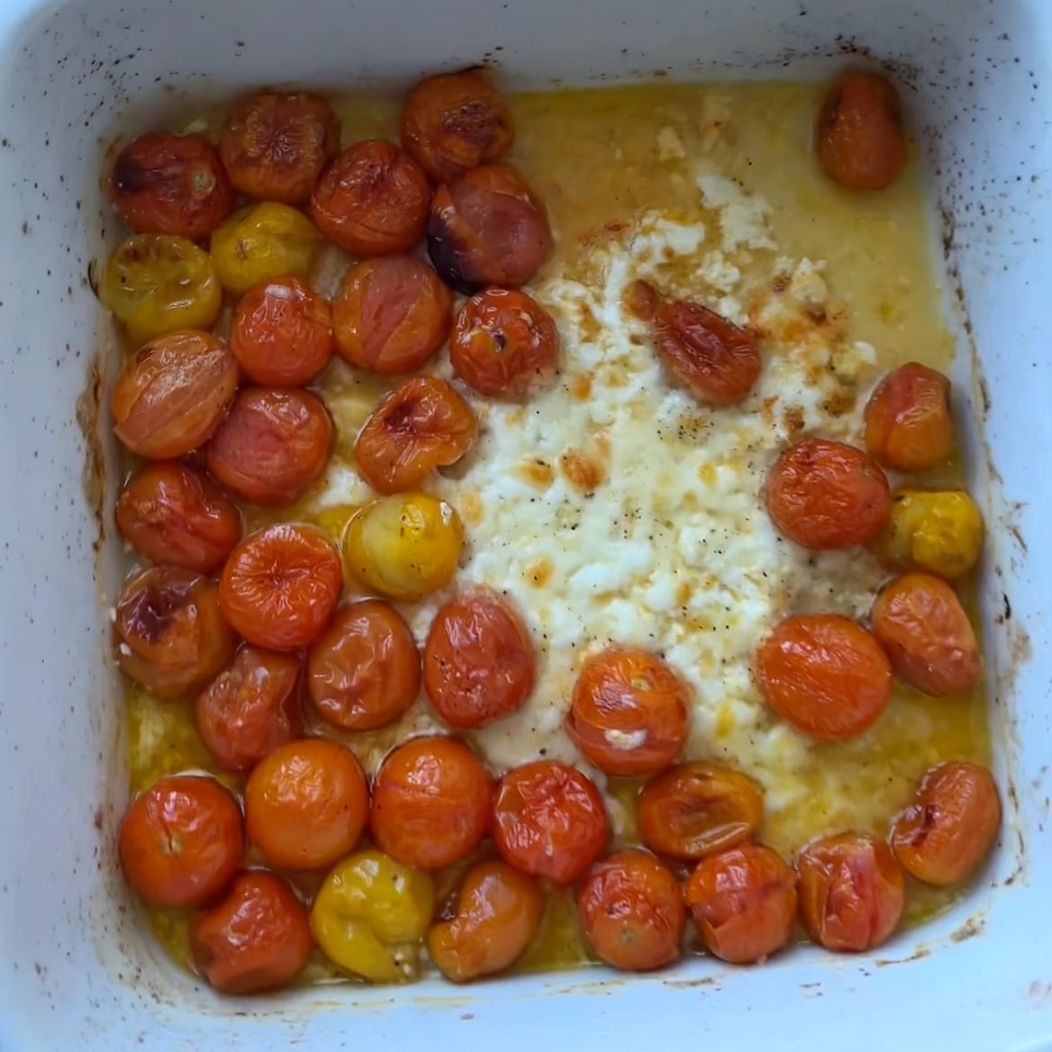 Roasted cherry tomatoes and a block of baked burrata cheese in a white baking dish, with olive oil and seasoning.