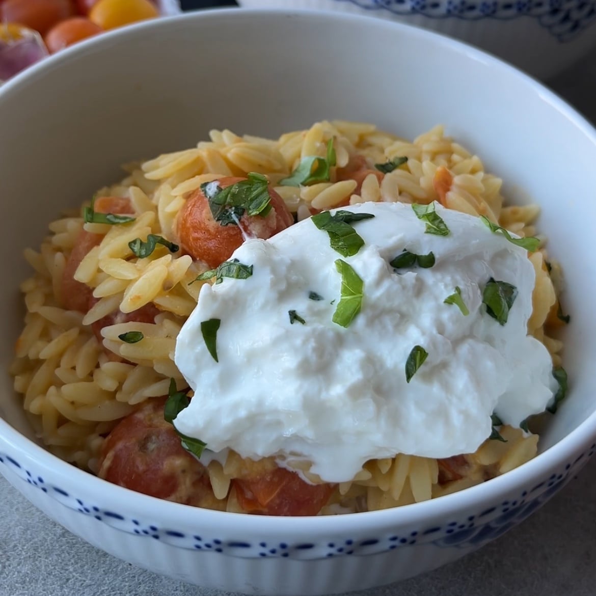 A bowl of orzo pasta with cherry tomatoes topped with a dollop of burrata and garnished with chopped herbs.
