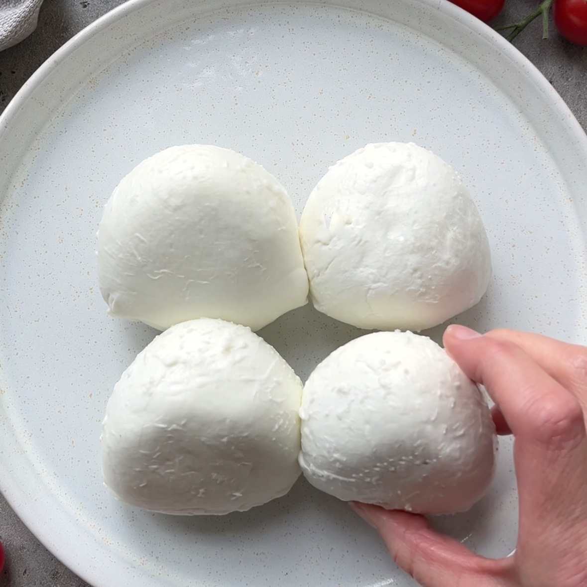 Four balls of fresh burrata cheese on a white plate, with a hand holding one piece in the lower right corner.