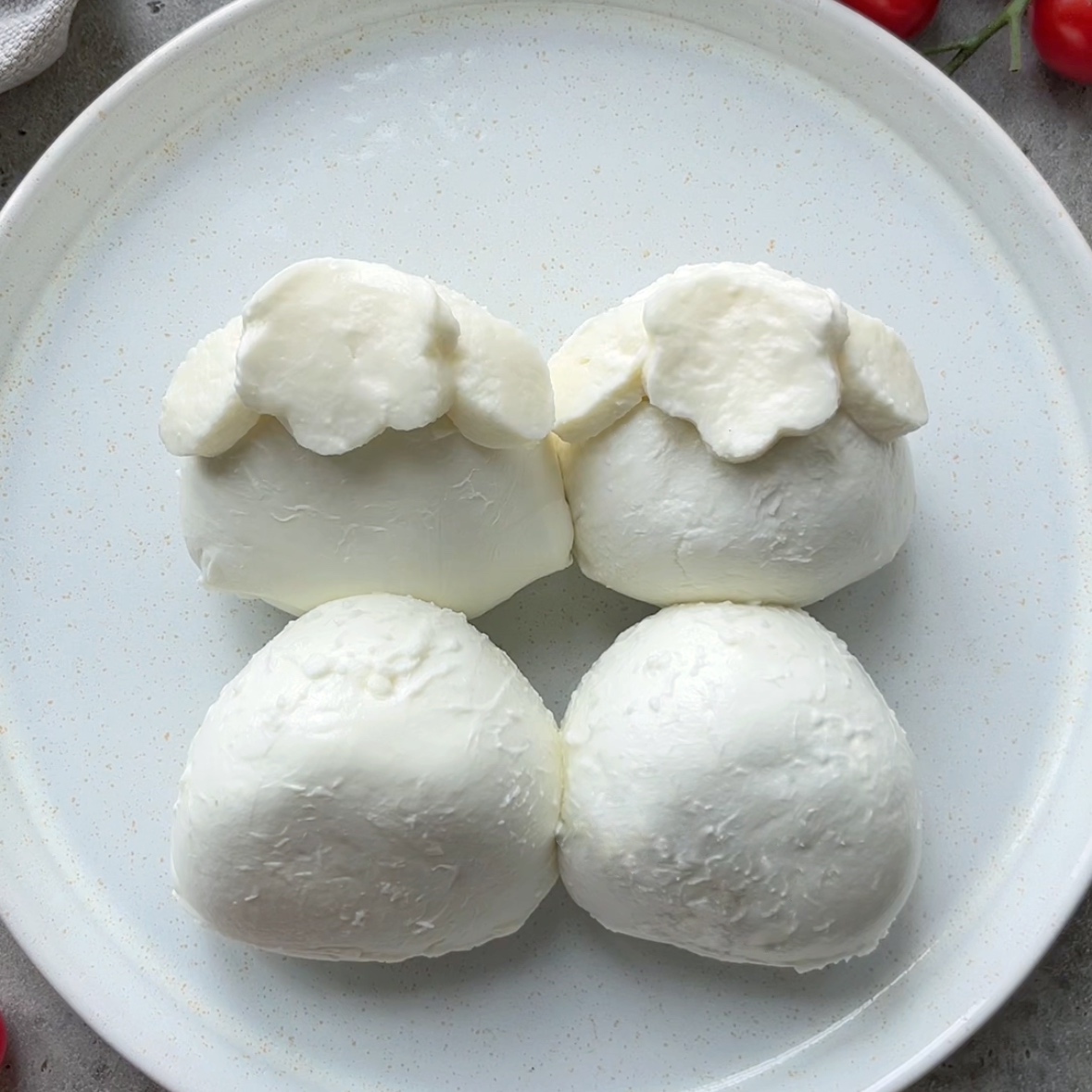 Four pieces of fresh burrata cheese are artfully arranged on a white plate, with two shaped to resemble lamb heads and two as their bodies.