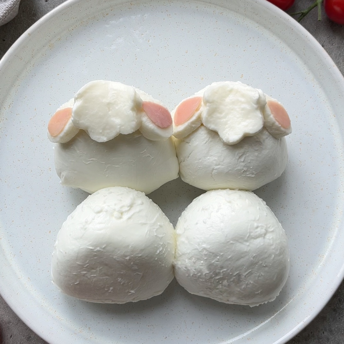Four balls of white cheese are arranged on a plate, with two decorated to resemble lamb faces using mozzarella and ham pieces.