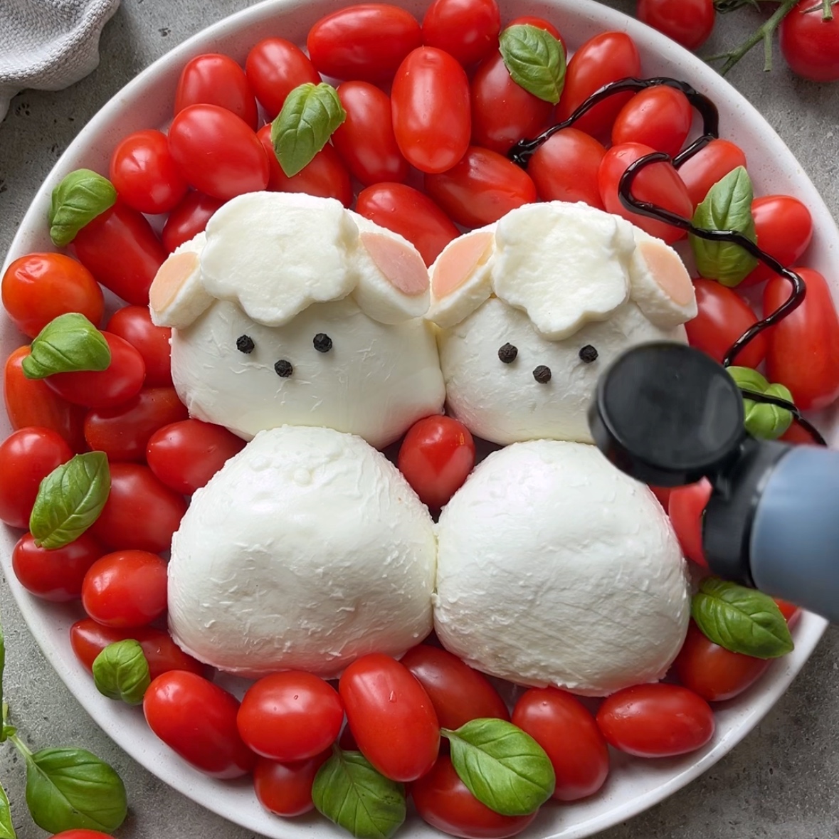 A plate with burrata shaped like two lamb, surrounded by grape tomatoes and basil leaves, with a hand pouring balsamic glaze from a bottle.