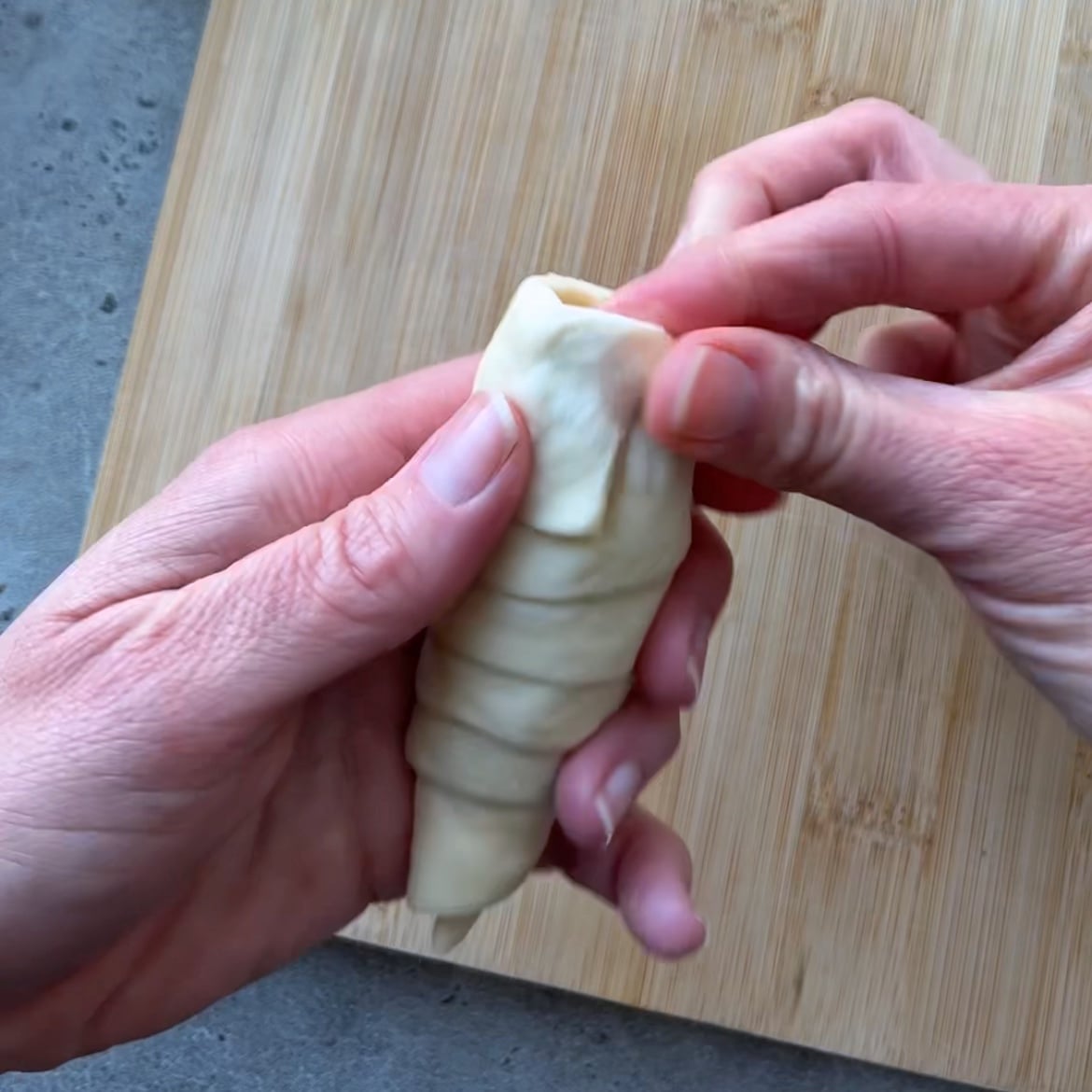 Two hands wrap dough around a pastry cone shape on a wooden cutting board.