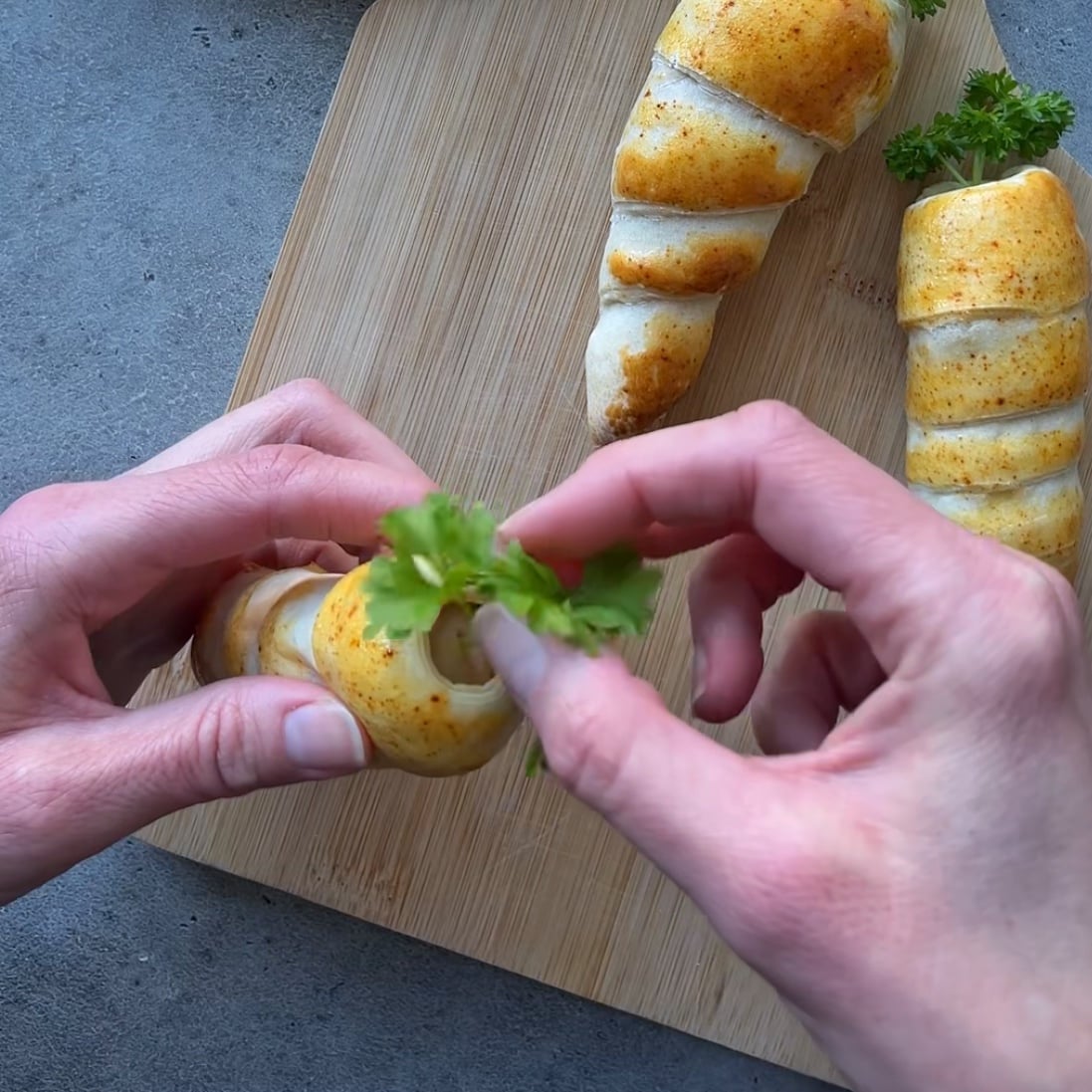Two hands place a sprig of parsley into a pastry shaped like a carrot, on a wooden cutting board.