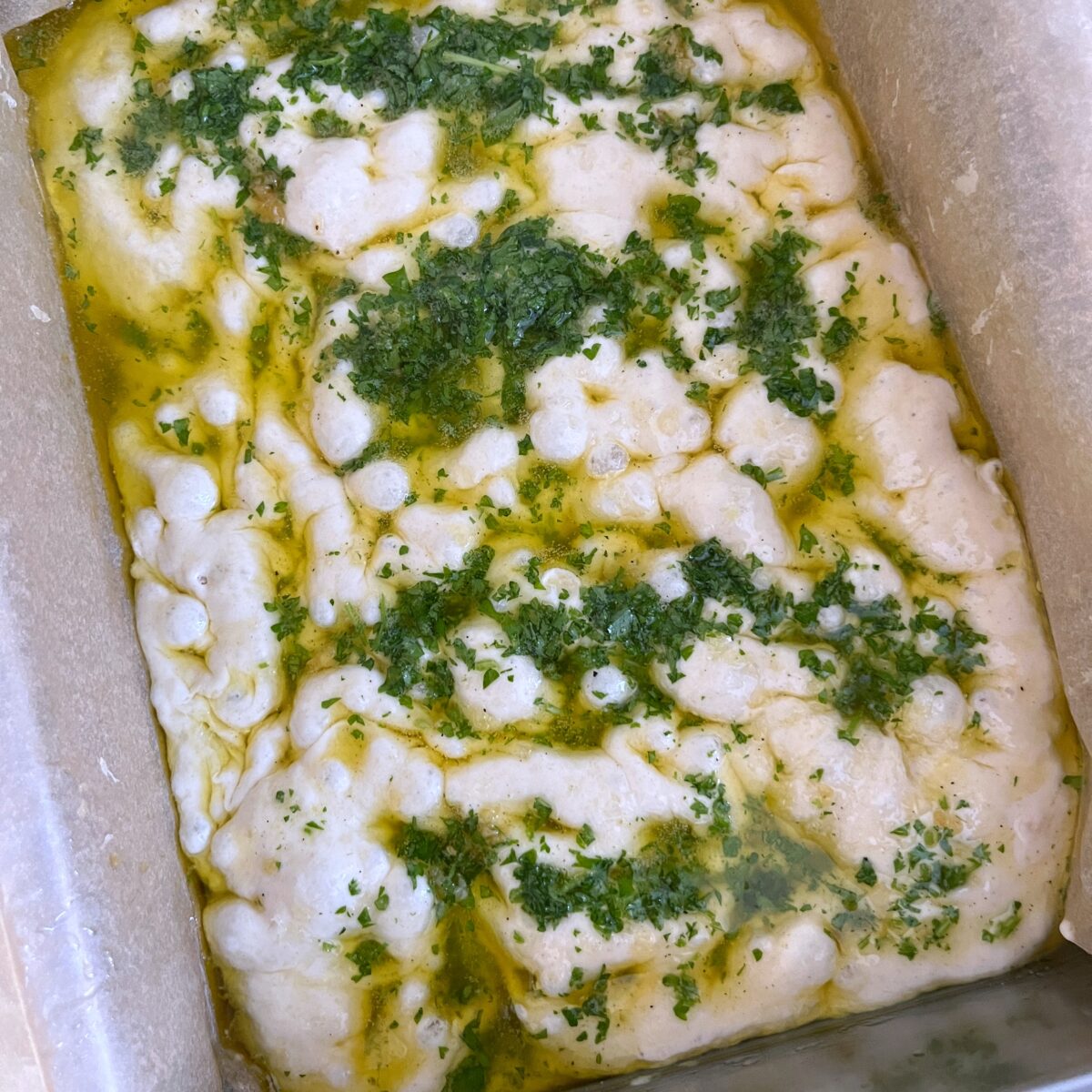 A close-up of unbaked Focaccia Garlic Bread dough in a parchment-lined pan, topped with olive oil and chopped herbs.