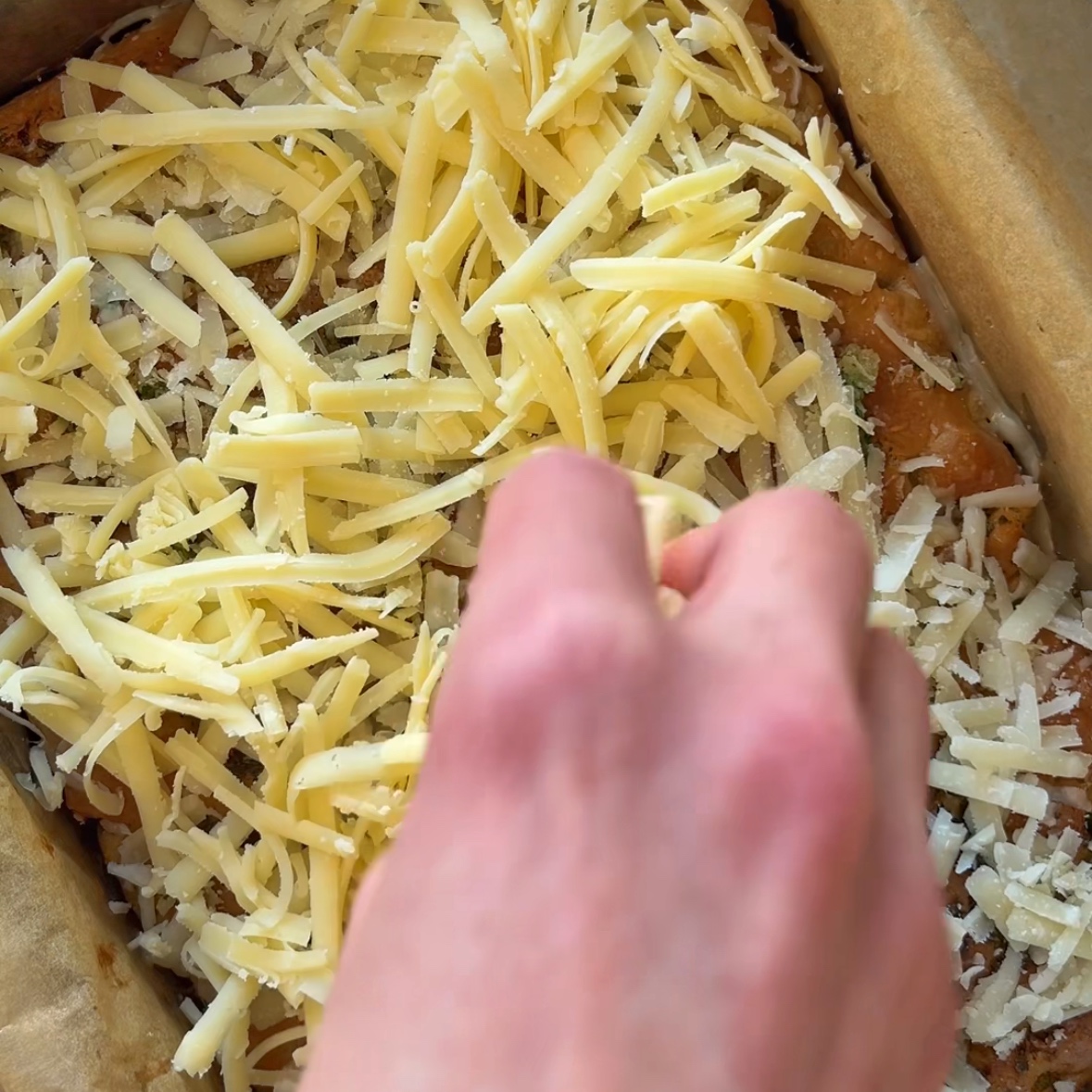A hand sprinkles grated cheddar on top of a baked focaccia, ready to be baked once more.