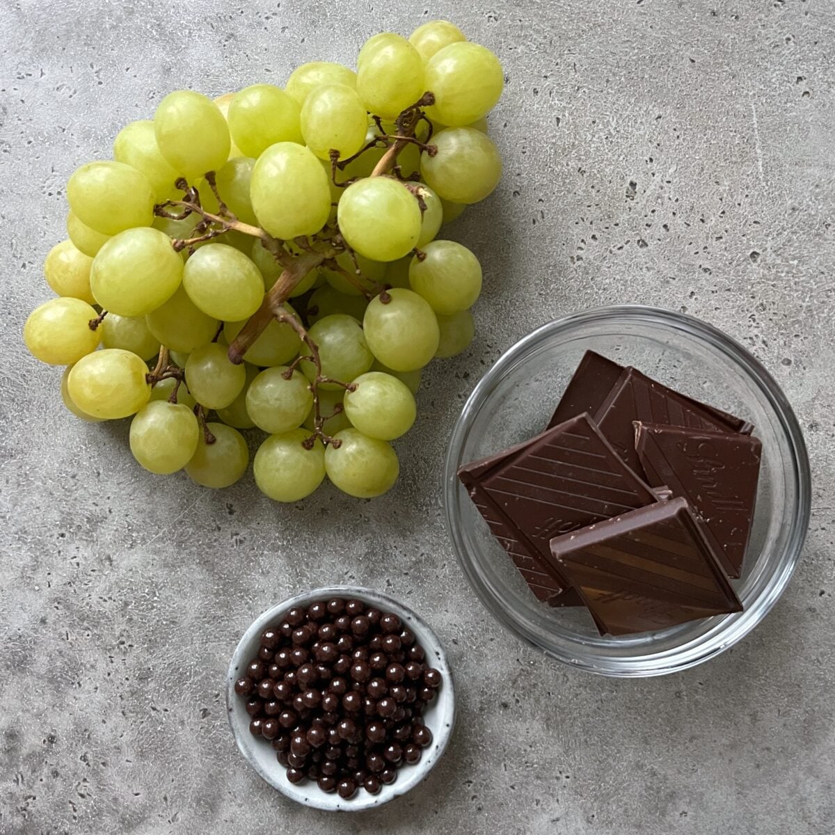 A bunch of green grapes, a glass bowl with chocolate squares, and a small dish with chocolate balls.
