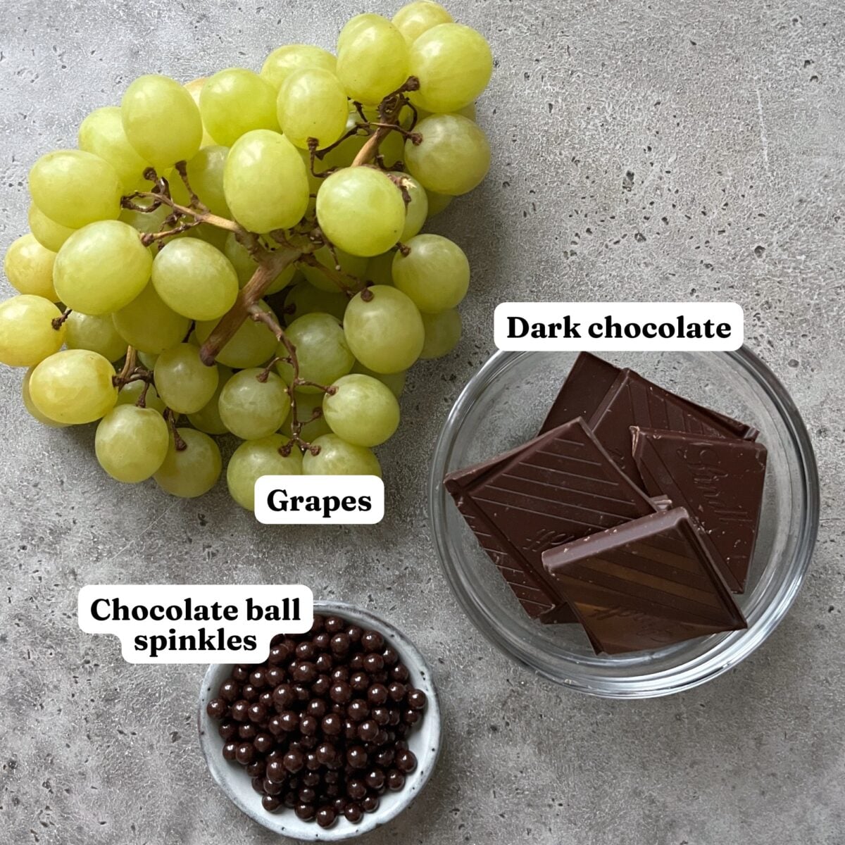 A bunch of green grapes, a bowl of dark chocolate pieces, and a small dish of chocolate sprinkles rest on a gray surface.