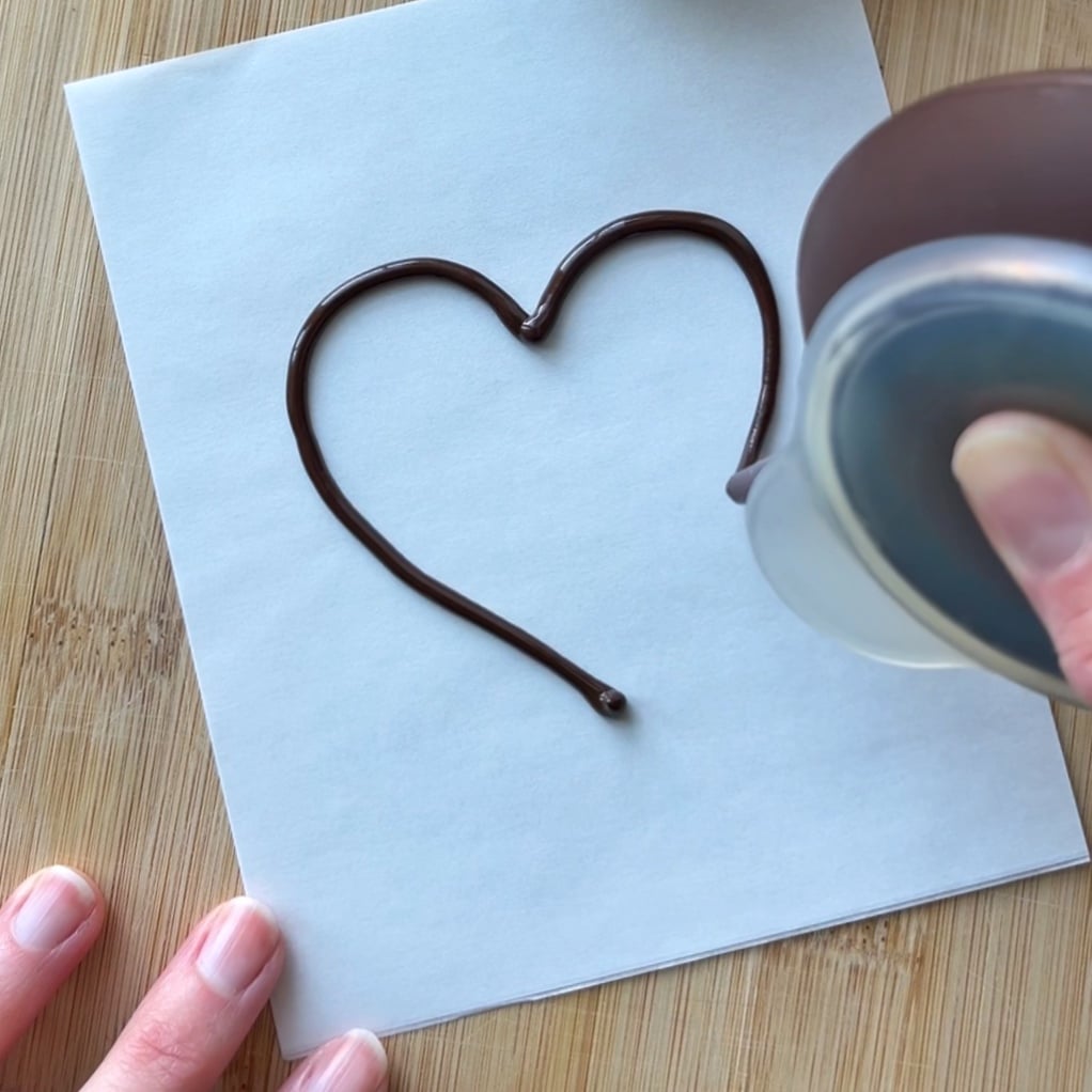 A hand pours chocolate in the shape of a heart onto a sheet of white parchment paper.