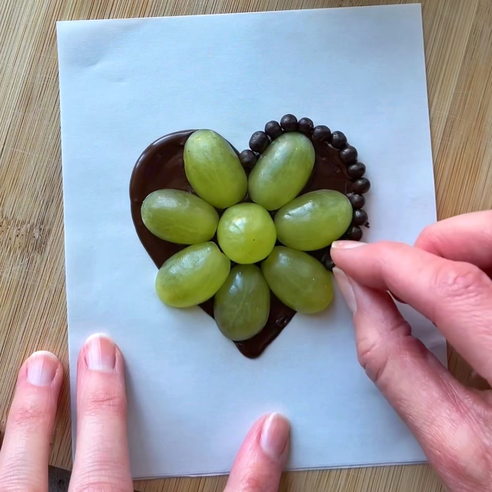 A hand decorates Grape Chocolate Hearts in a heart-shaped arrangement of green grapes and chocolate balls.