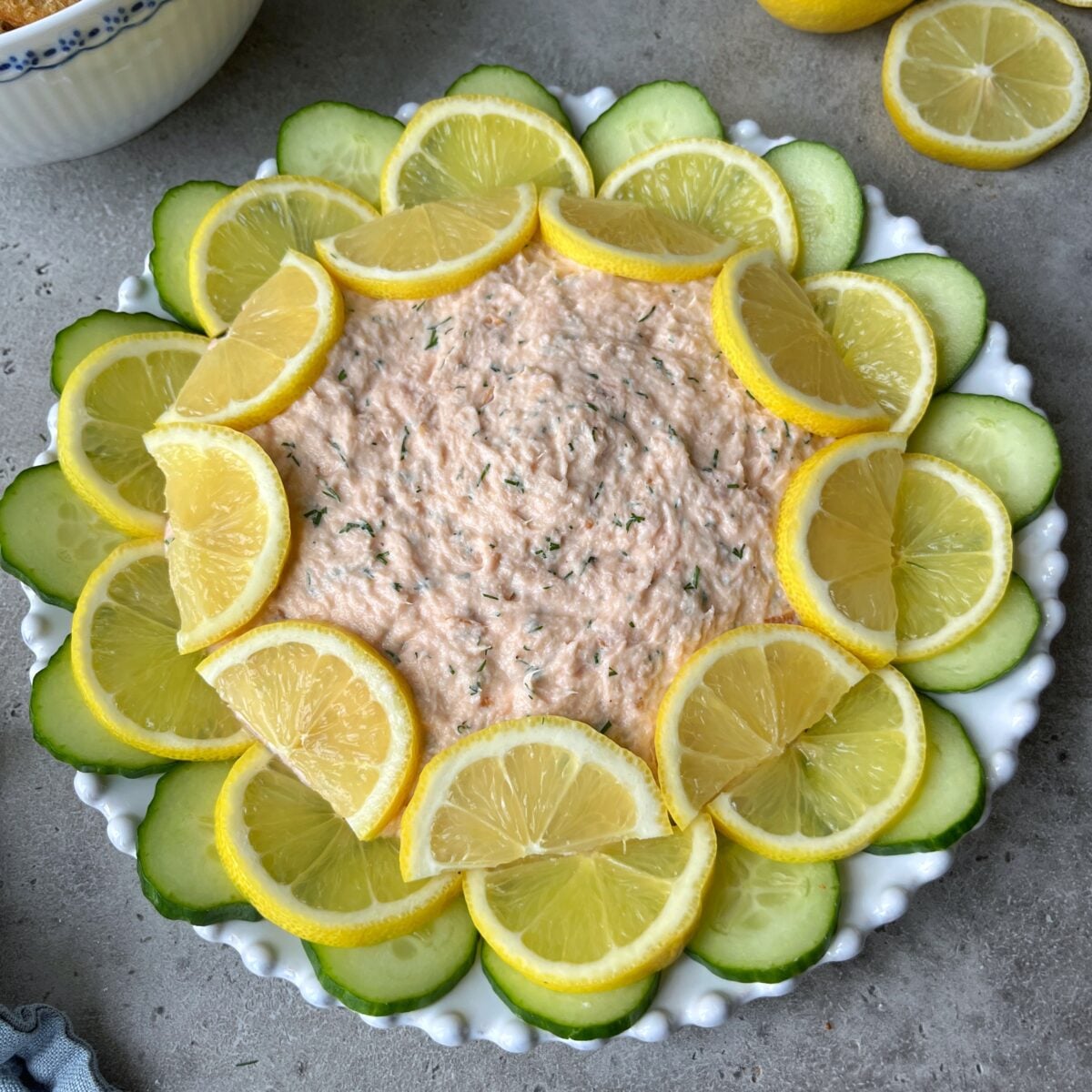 A round dish of Hot Smoked Salmon Dip topped with dill, surrounded by alternating slices of lemon and cucumber, on a white plate.
