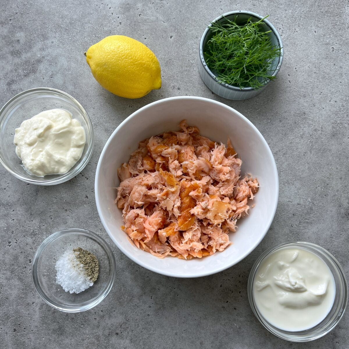 A bowl of flaked salmon, Greek yogurt, fresh dill, salt and pepper, a lemon, and creme fraiche on a gray surface.