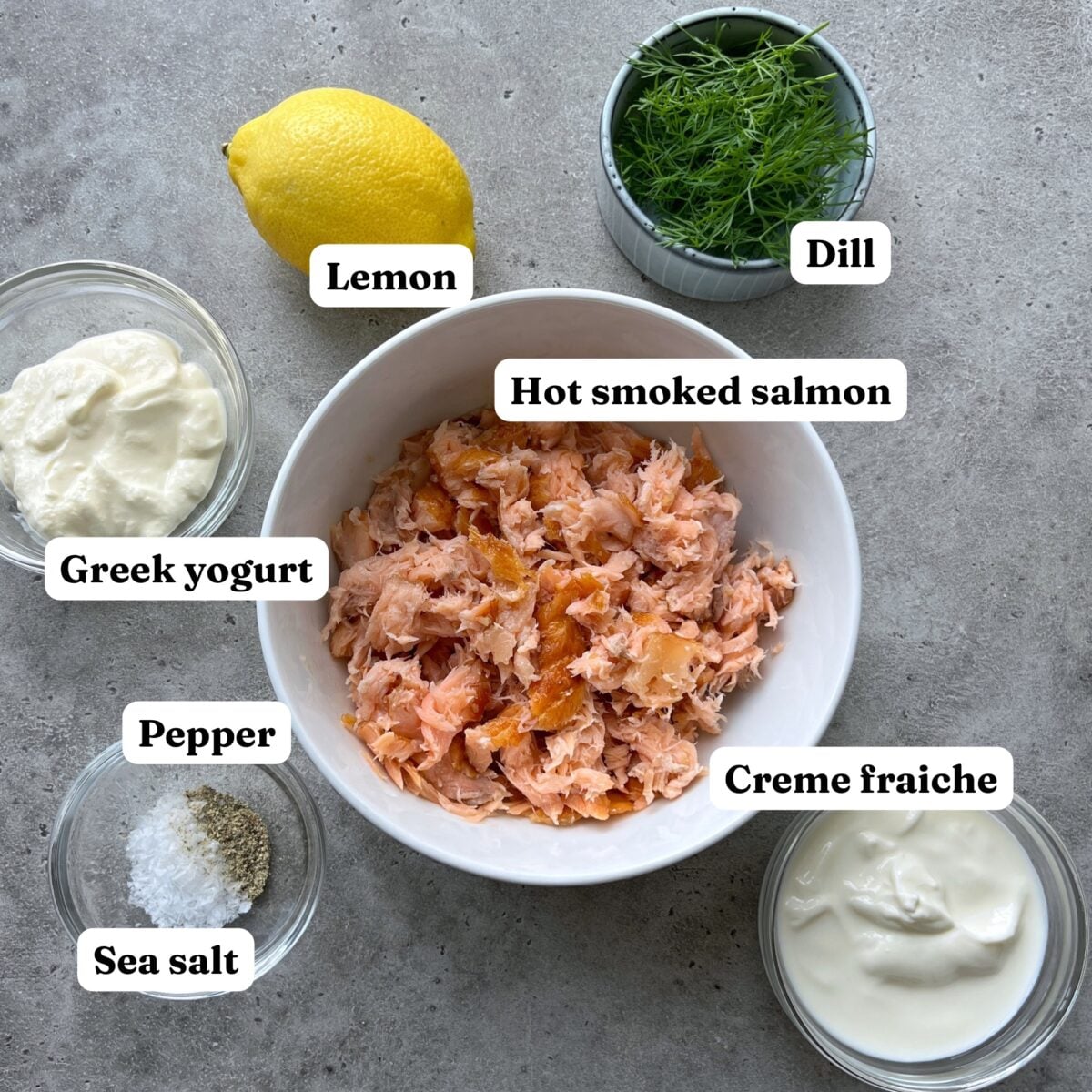 A bowl of flaked hot smoked salmon is surrounded by labeled ingredients including lemon, dill, Greek yogurt, crรจme fraรฎche, sea salt, and pepper on a gray surface.