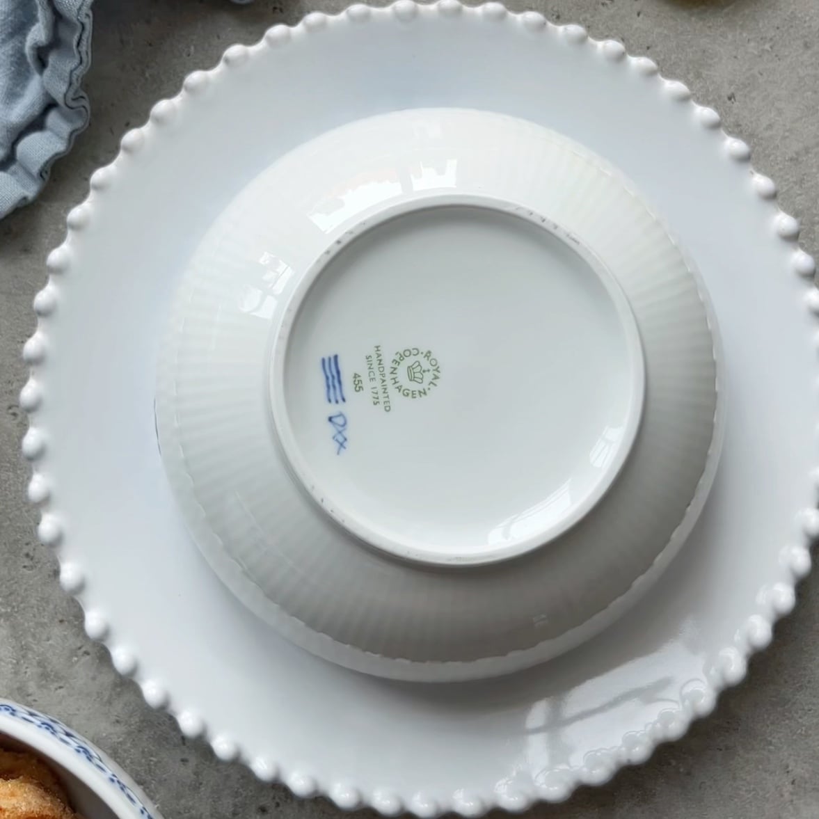 A white ceramic bowl is facing down on top of a white plate with a beaded rim, set on a gray surface.