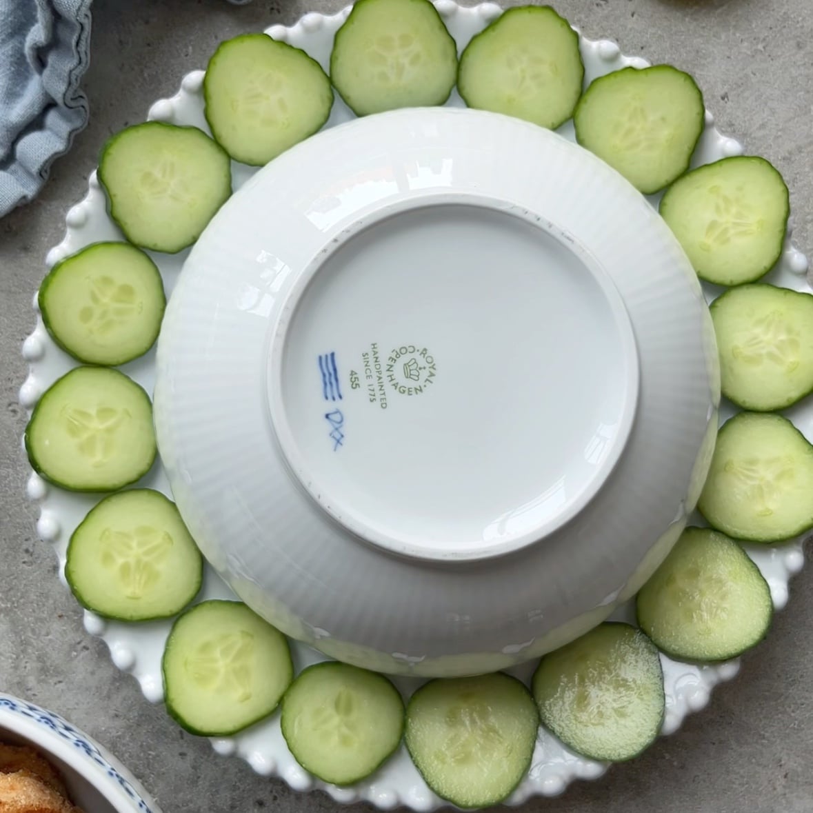 A white plate is arranged with cucumber slices around the edge, with an upside-down white bowl placed in the center.
