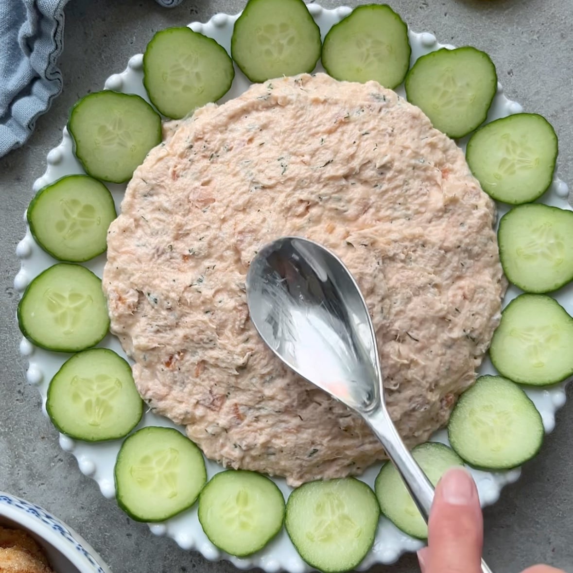 A hand spreads a creamy Hot Smoked Salmon Dip on a plate, surrounded by sliced cucumber rounds.