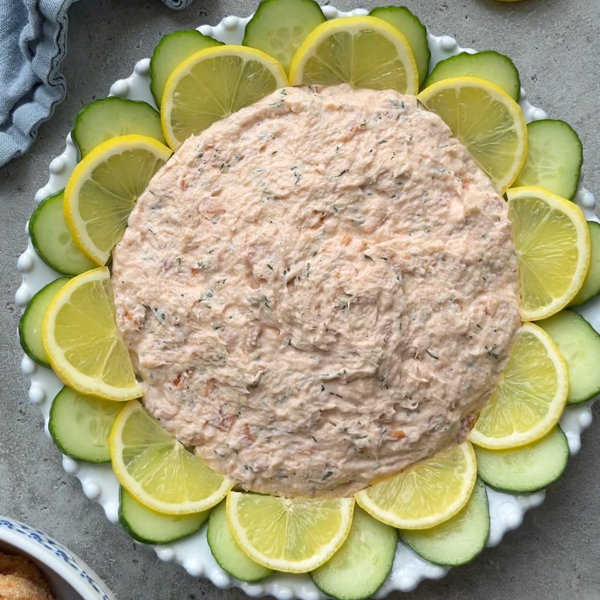 A round plate of Hot Smoked Salmon Dip is surrounded by alternating slices of cucumber and lemon, arranged in a circular pattern on a textured gray surface.