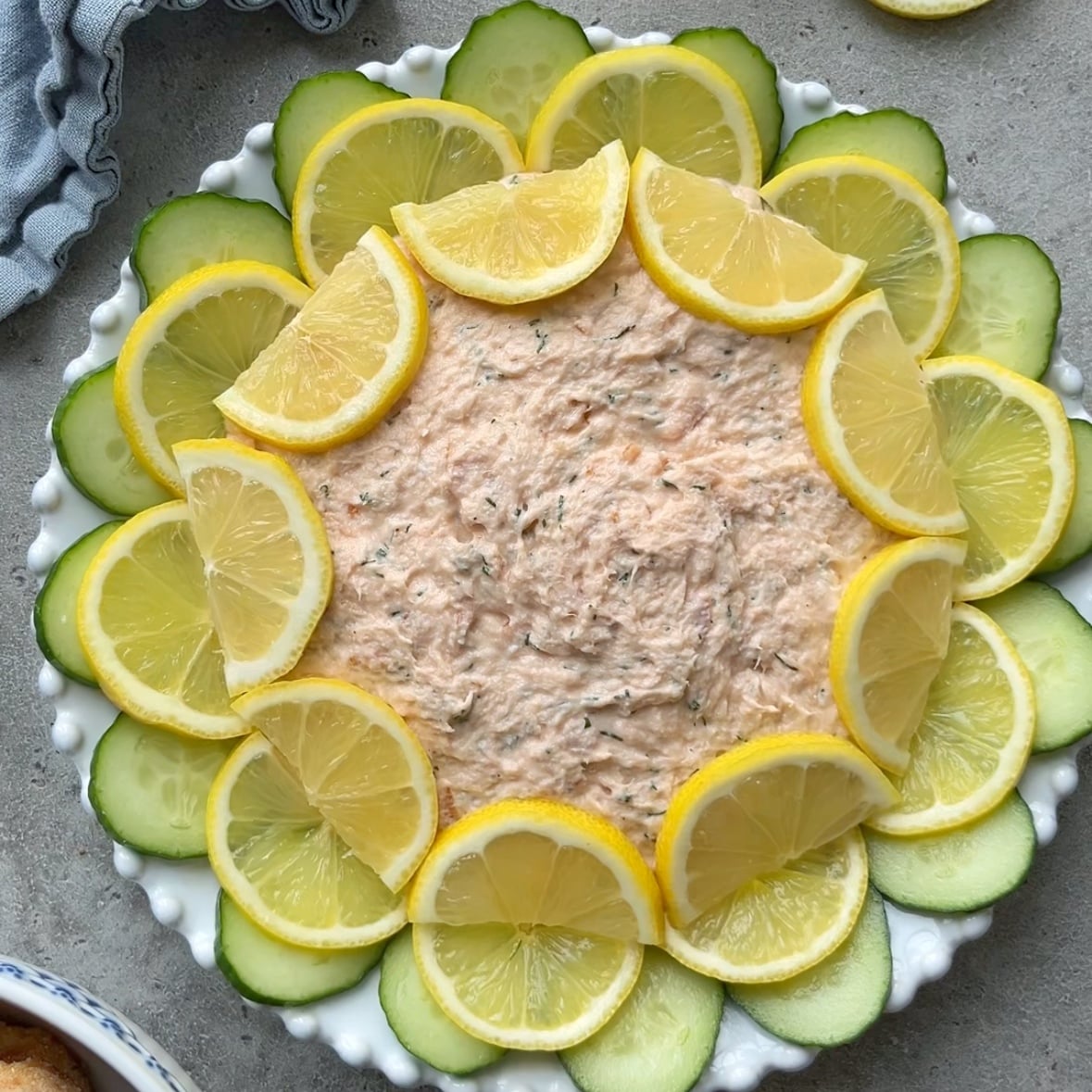 A dish of Hot Smoked Salmon Dip is garnished with overlapping slices of lemon and cucumber arranged in a circular pattern on a white plate.