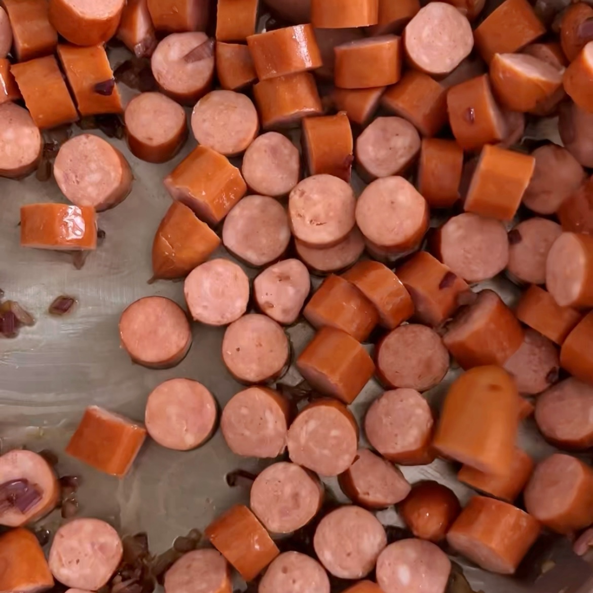 Sliced cooked sausage pieces in a pan, with some small bits of chopped onions visible among them.